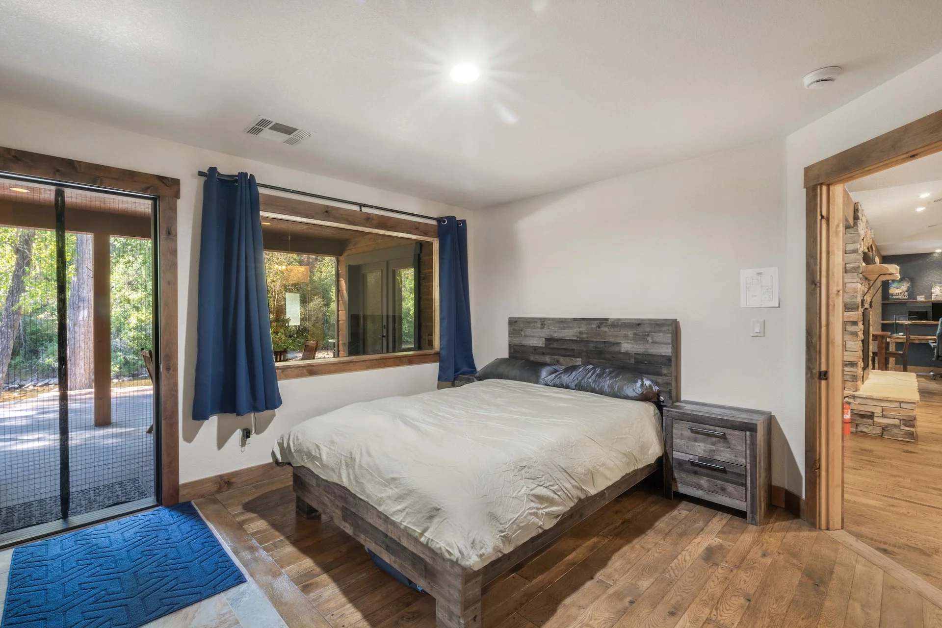 A bedroom with a wooden bedframe and nightstand, beige bedding, a window with blue curtains, a door to a outdoor patio, and a wooden door frame leading to another room.