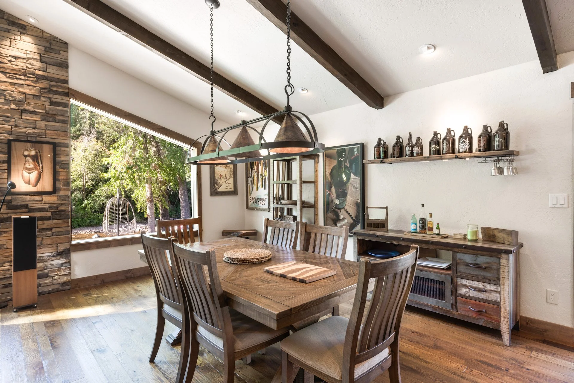 A rustic dining room with a wooden table and chairs, a large window showing trees outside, and a wooden sideboard with various bottles and decorations, decorated with artwork and a hanging lamp.