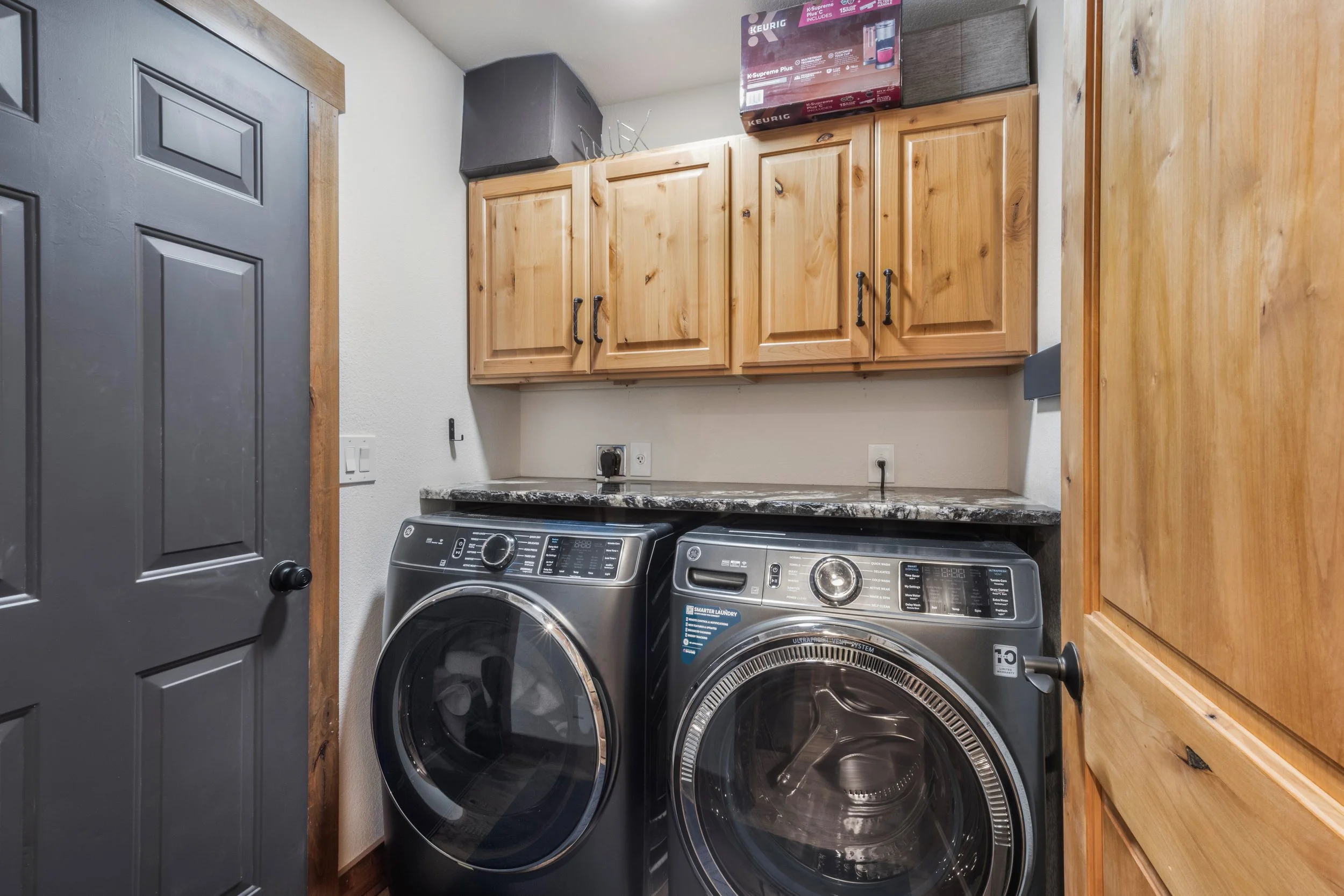 A laundry room with a washer and dryer side by side, wooden cabinets above, and a gray door on the left. Items are stored on top of the cabinets.