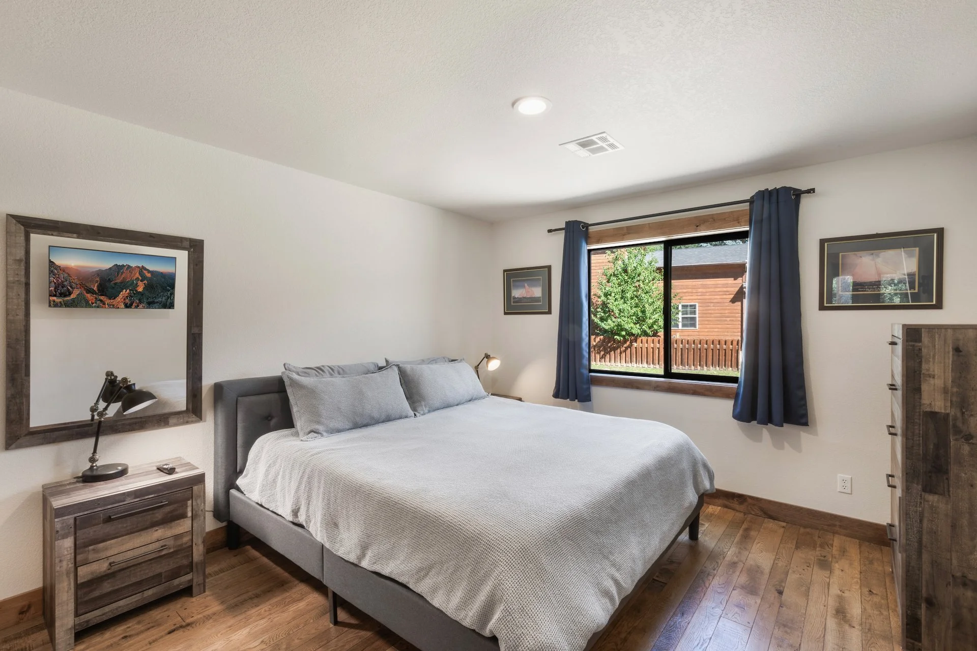 A bedroom with a large bed covered in gray bedding and pillows, a wooden nightstand with a desk lamp, and a window with blue curtains showing a view of a wooden fence and greenery outside.
