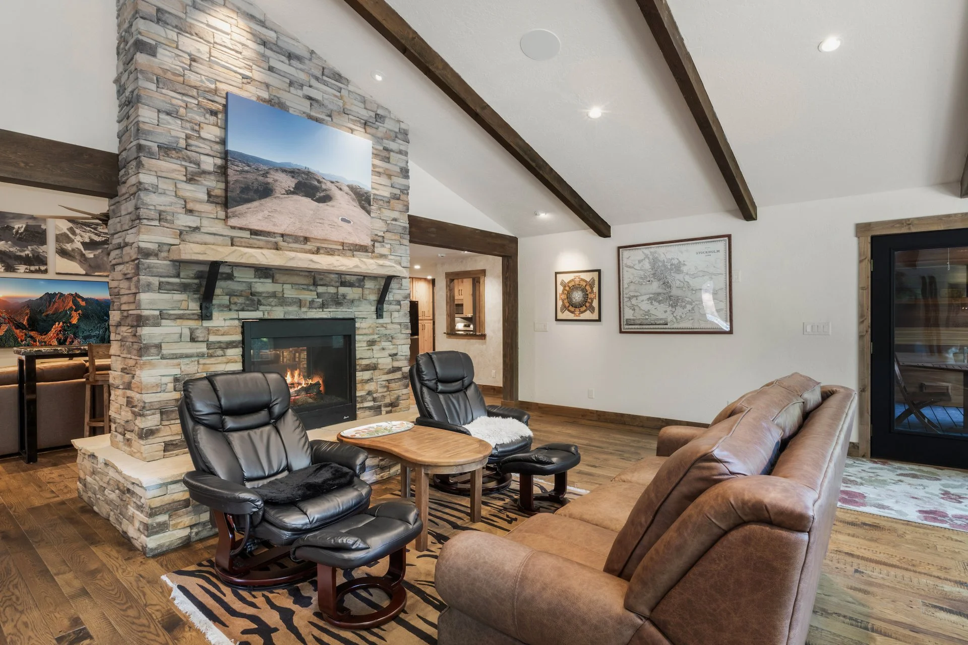 Living room with a stone fireplace, black leather armchairs, a brown leather couch, and framed artwork on white walls with dark wooden ceiling beams.