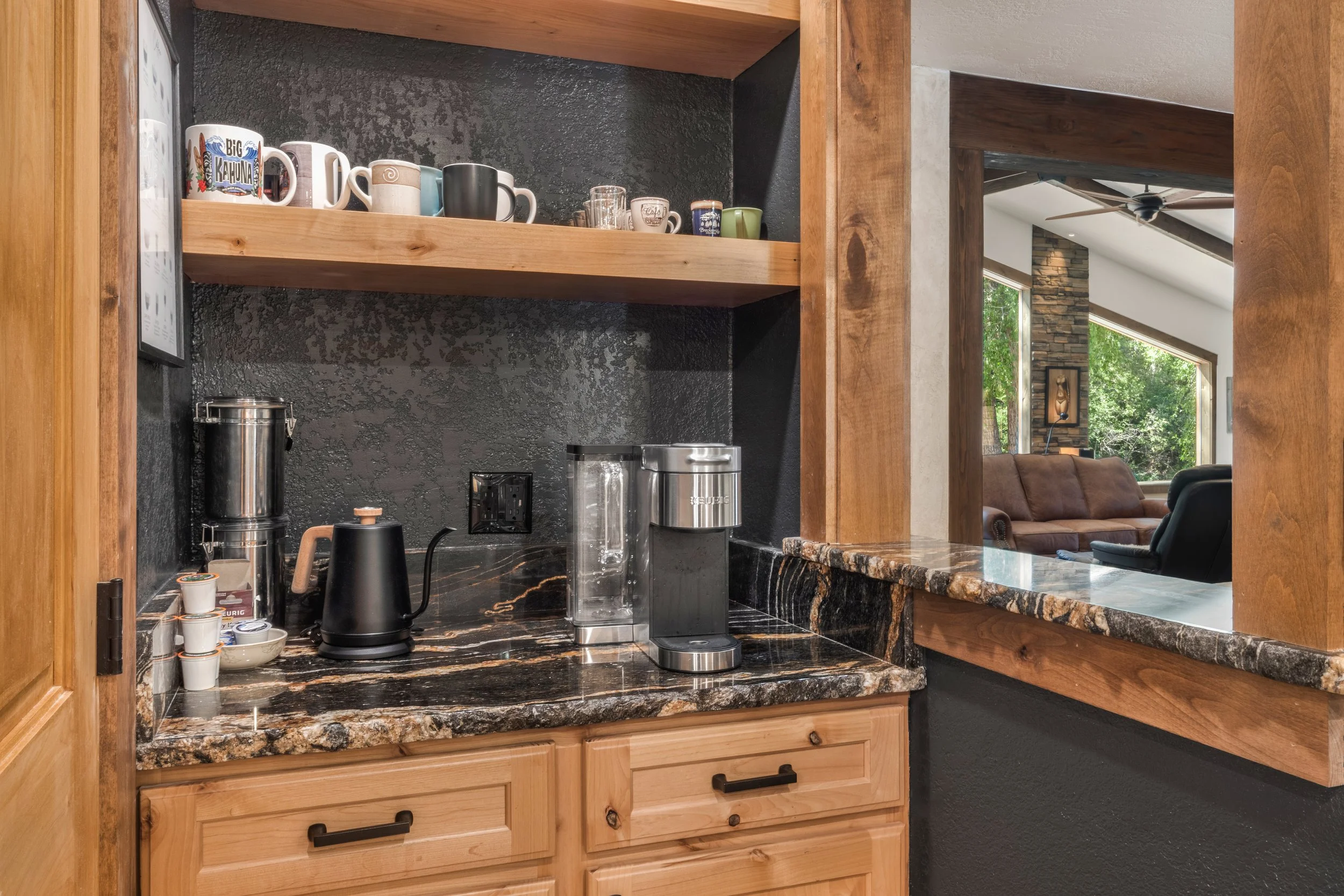 Kitchen counter with coffee maker, water filter, kettle, and coffee pods, open shelves with mugs and cups, view into living room with brown sofa and large windows.
