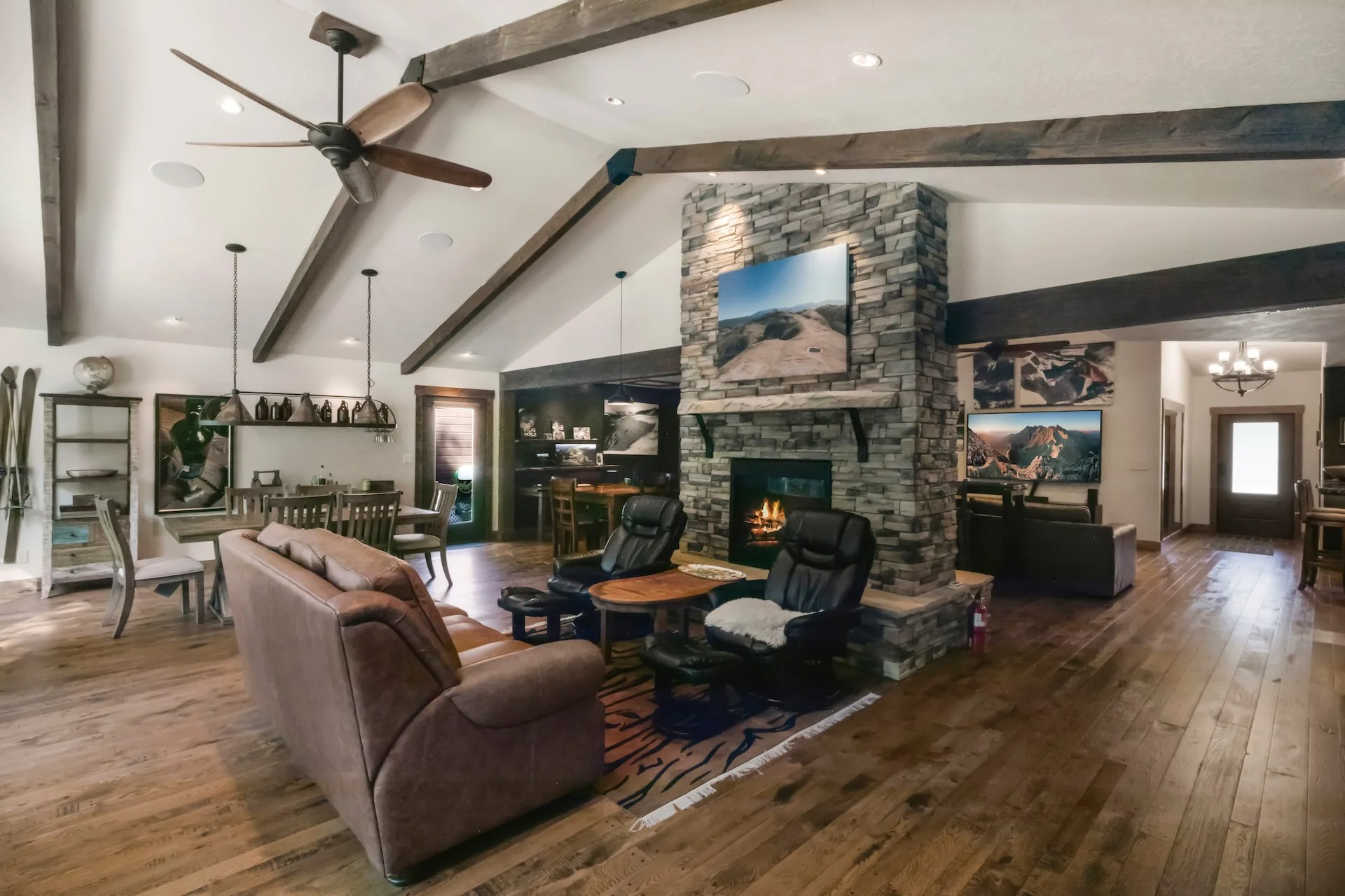 Spacious living room with wooden floors, stone fireplace, leather couch, black chairs, dining table, and mounted TV surrounded by wooden beams on the ceiling.