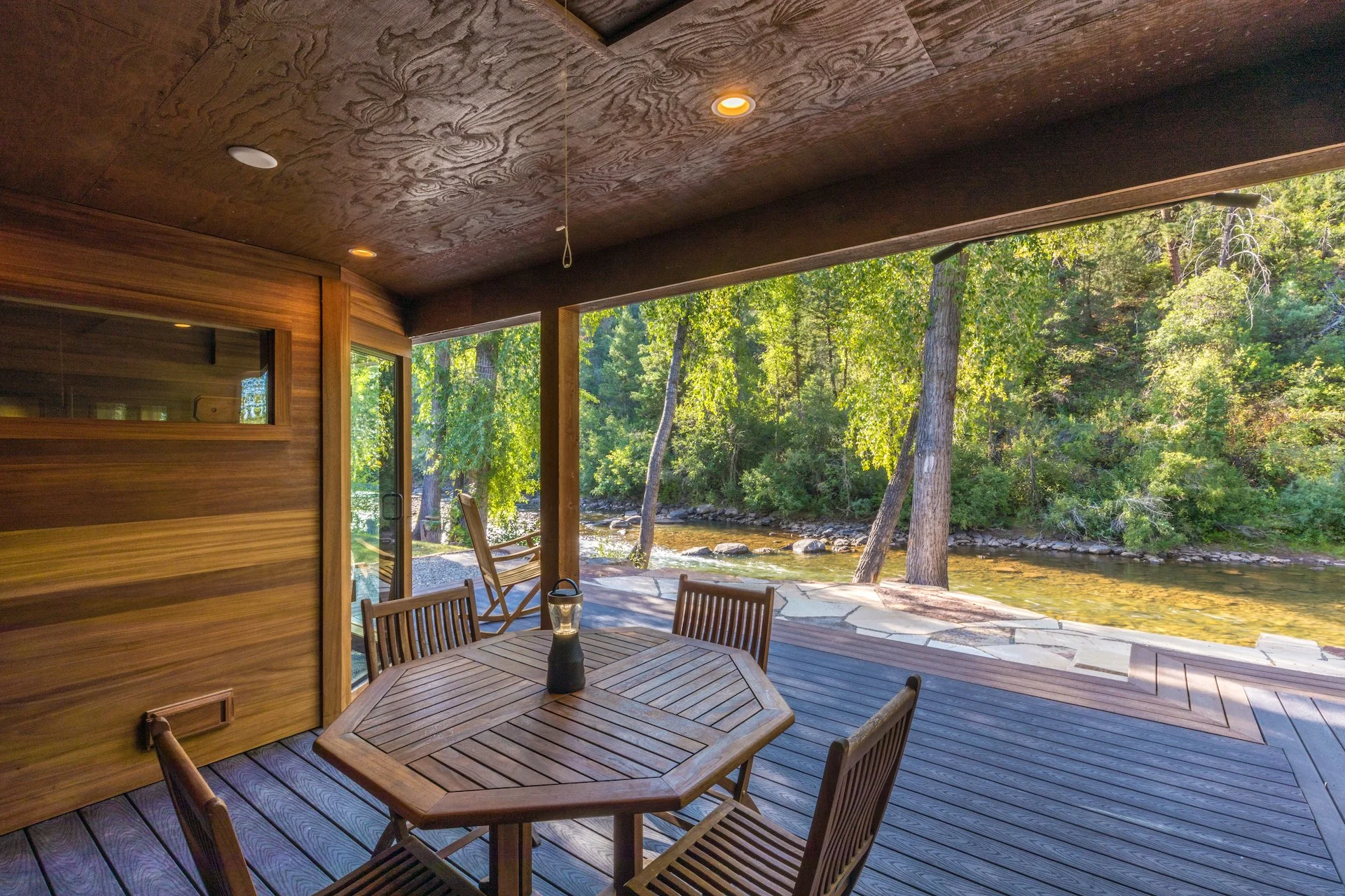 A covered wooden deck with a round table and four chairs overlooks a river with trees in the background.
