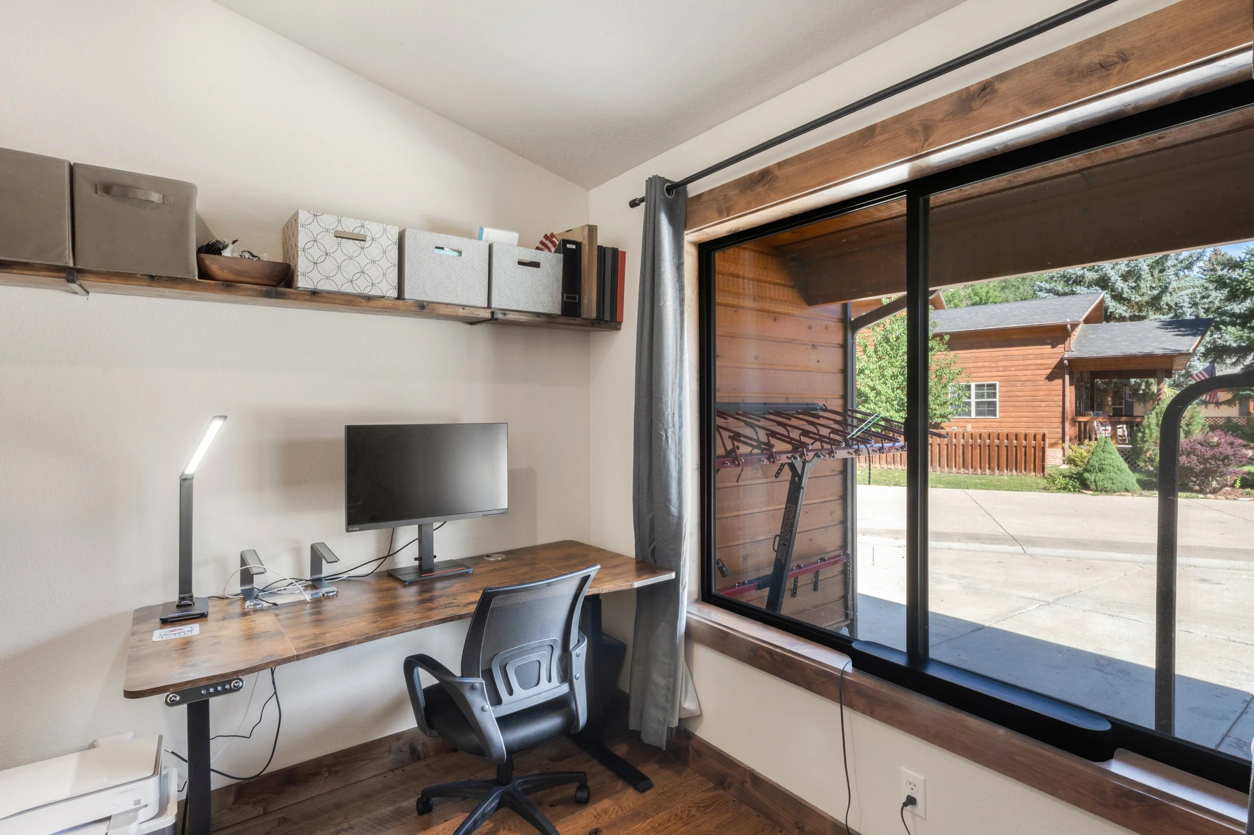 Home office with a wooden desk, a computer monitor, a black office chair, a desk lamp, and shelves on the wall holding storage boxes and binders. Large window with a curtain, overlooking a backyard with a clothes drying rack, trees, and neighboring h