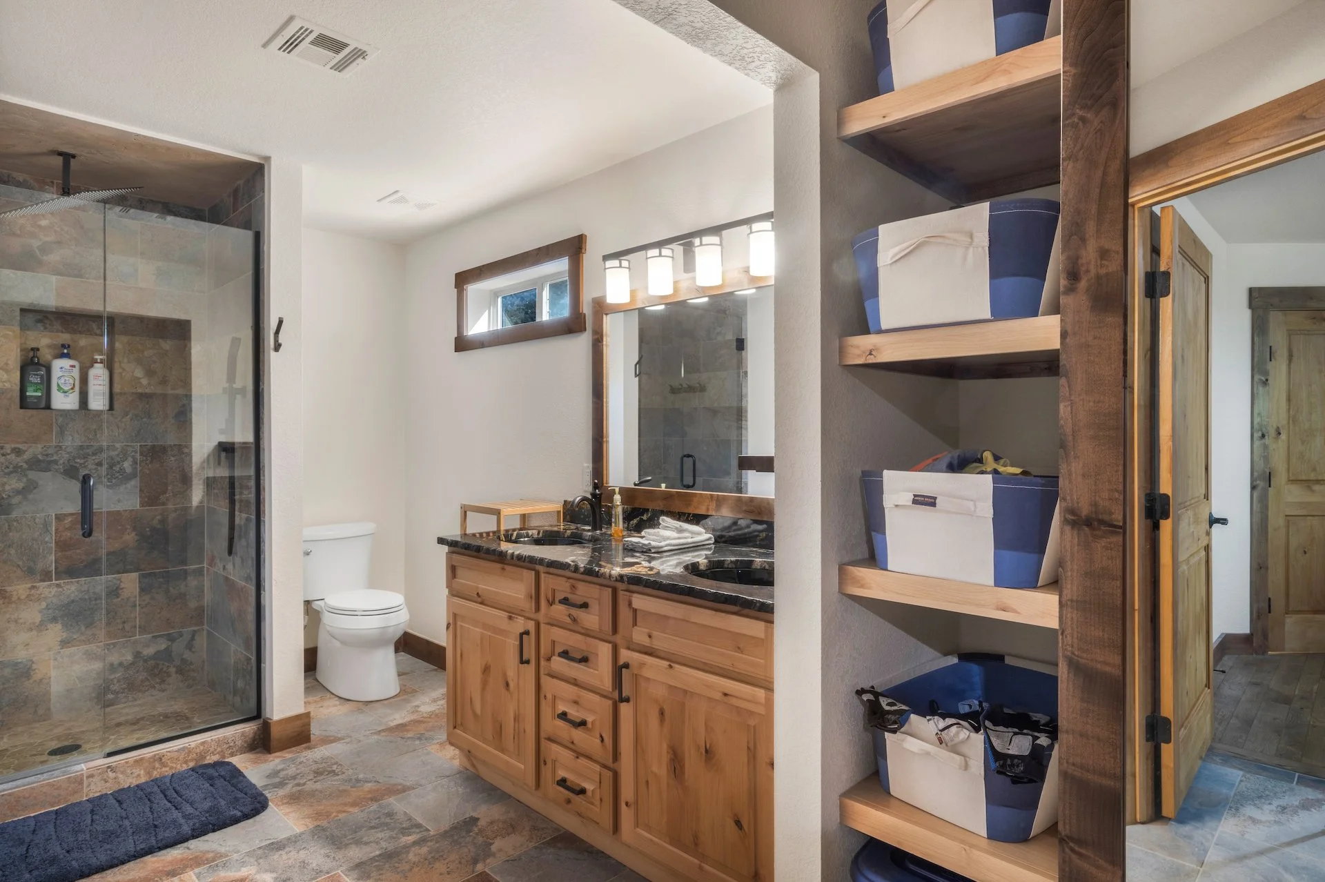 Bathroom with walk-in shower, toilet, wooden vanity with black granite countertop, mirror, wall-mounted lights, small window, and open shelves with storage bins.