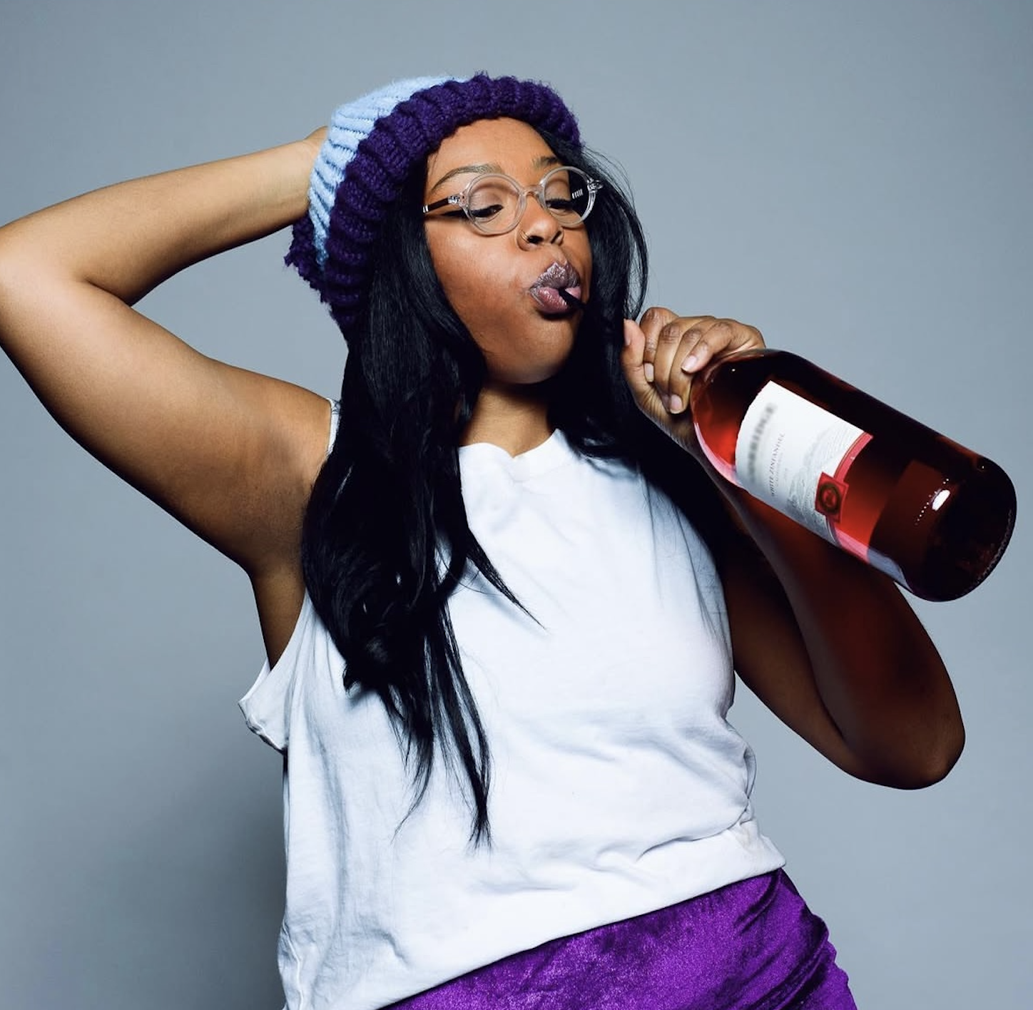 Rachel Hall playing Mara, co-star of Not Flexible, wearing glasses and a purple and blue knit beanie holding a bottle of rosé wine, appearing to pretend drink from it