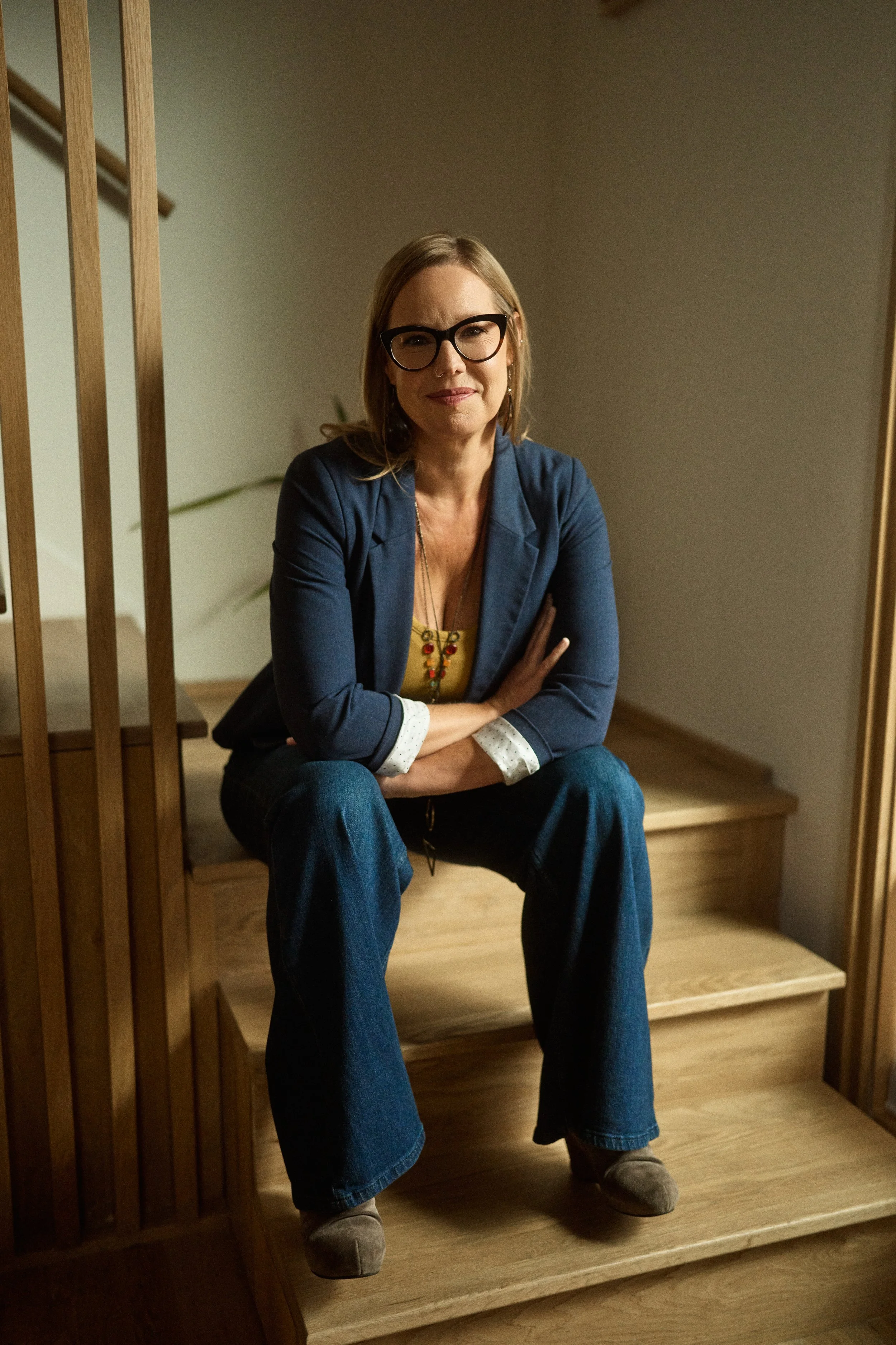 A woman with blonde hair and glasses sitting on wooden stairs, arms crossed, looking at the camera in a cozy indoor setting.