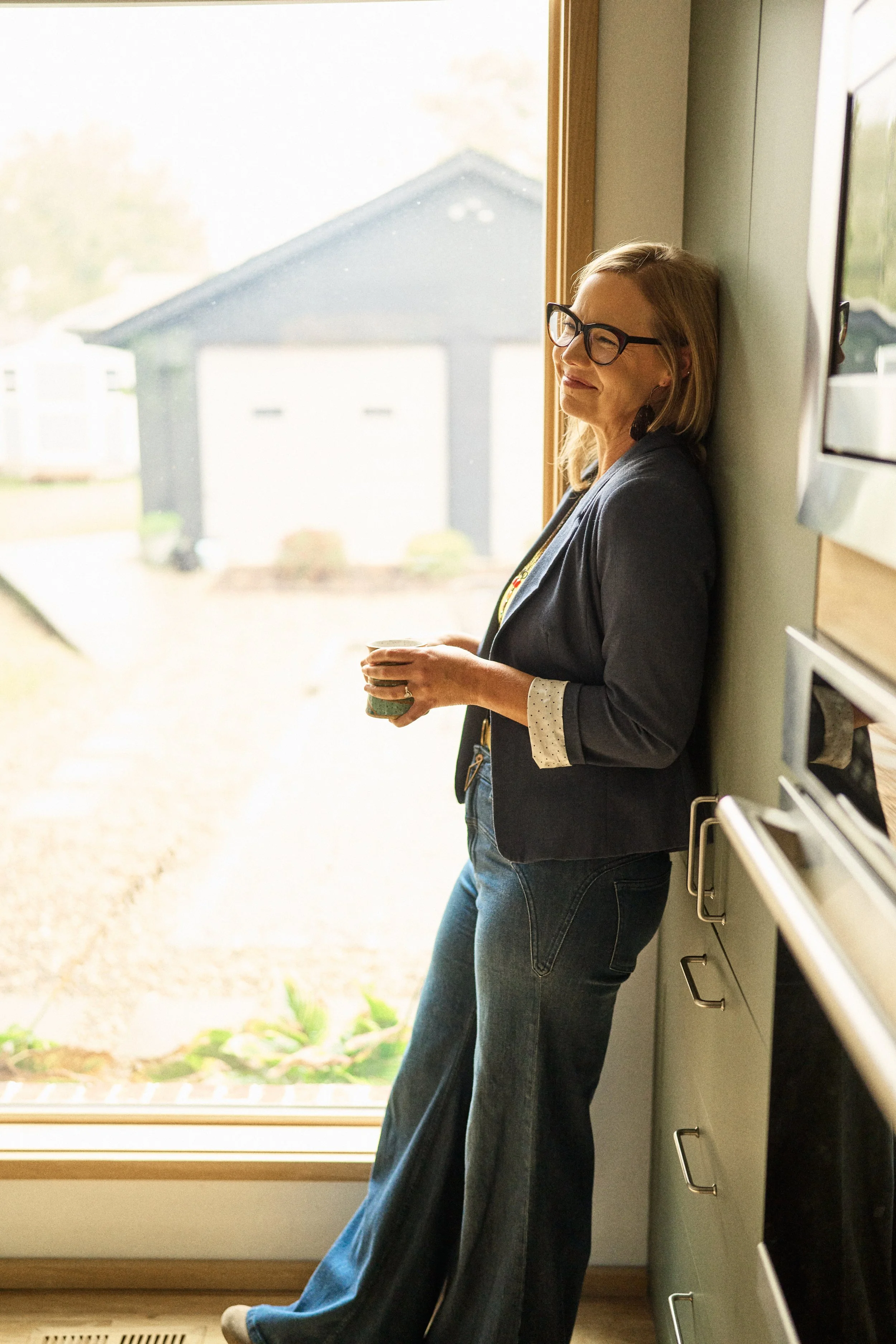 A woman with glasses and shoulder-length blonde hair leaning against a wall, holding a cup, looking out a large window with a house and yard outside.