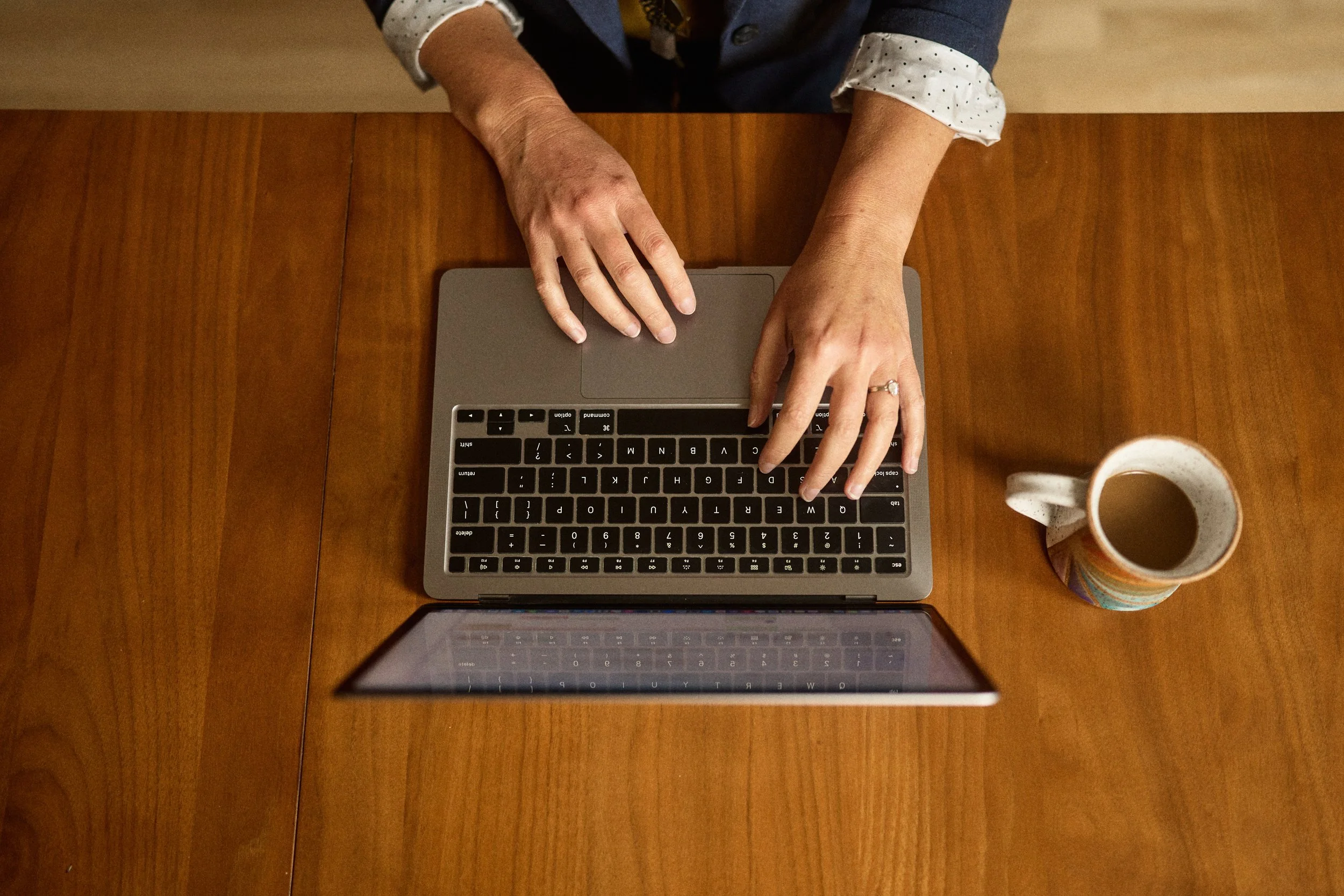 Top view of a person's hands typing on a laptop keyboard on a wooden desk, with a coffee mug to the side.