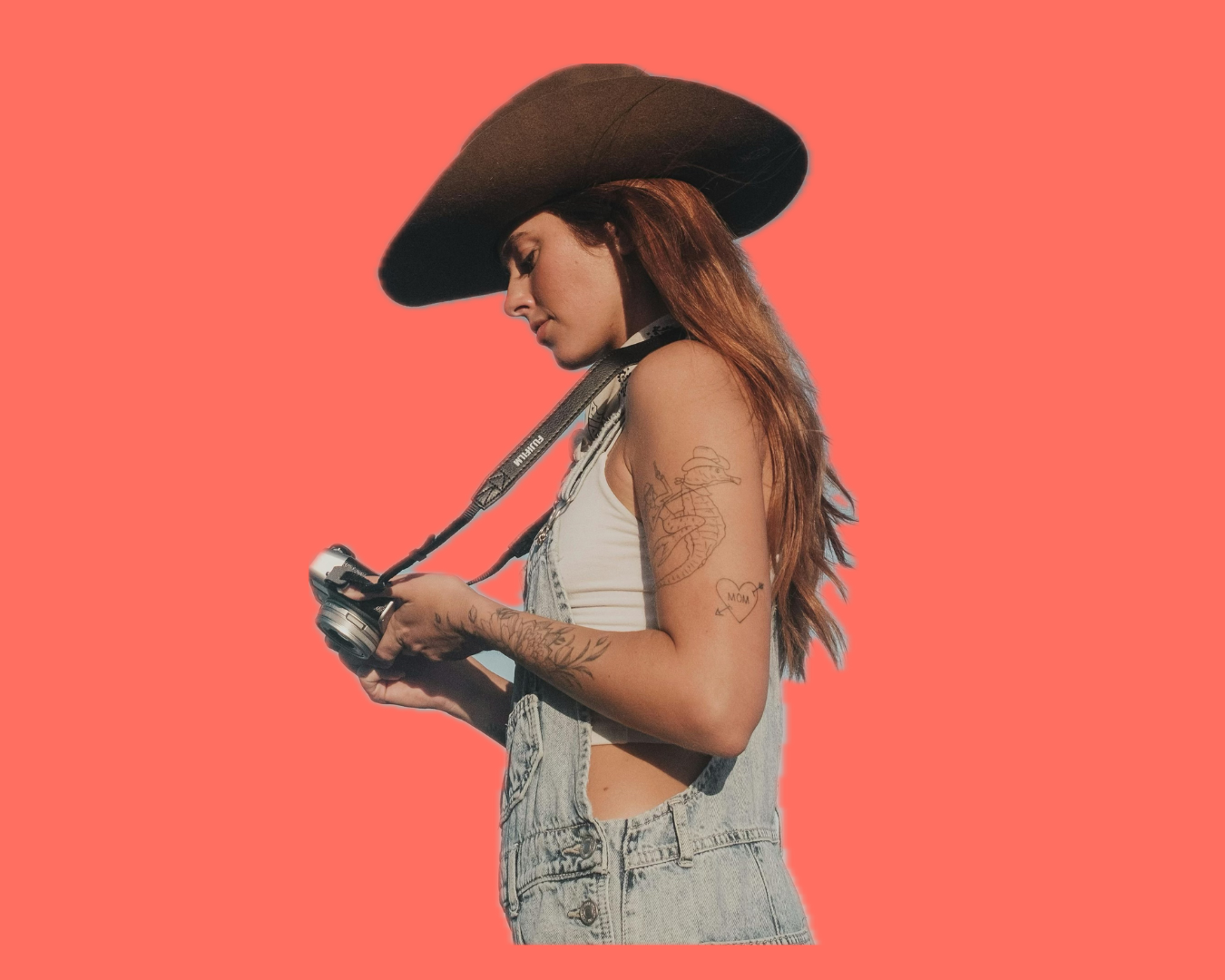 A young woman with long hair wearing a black wide-brimmed hat, white tank top, and denim overalls, looking at a camera she is holding, set against a coral background.