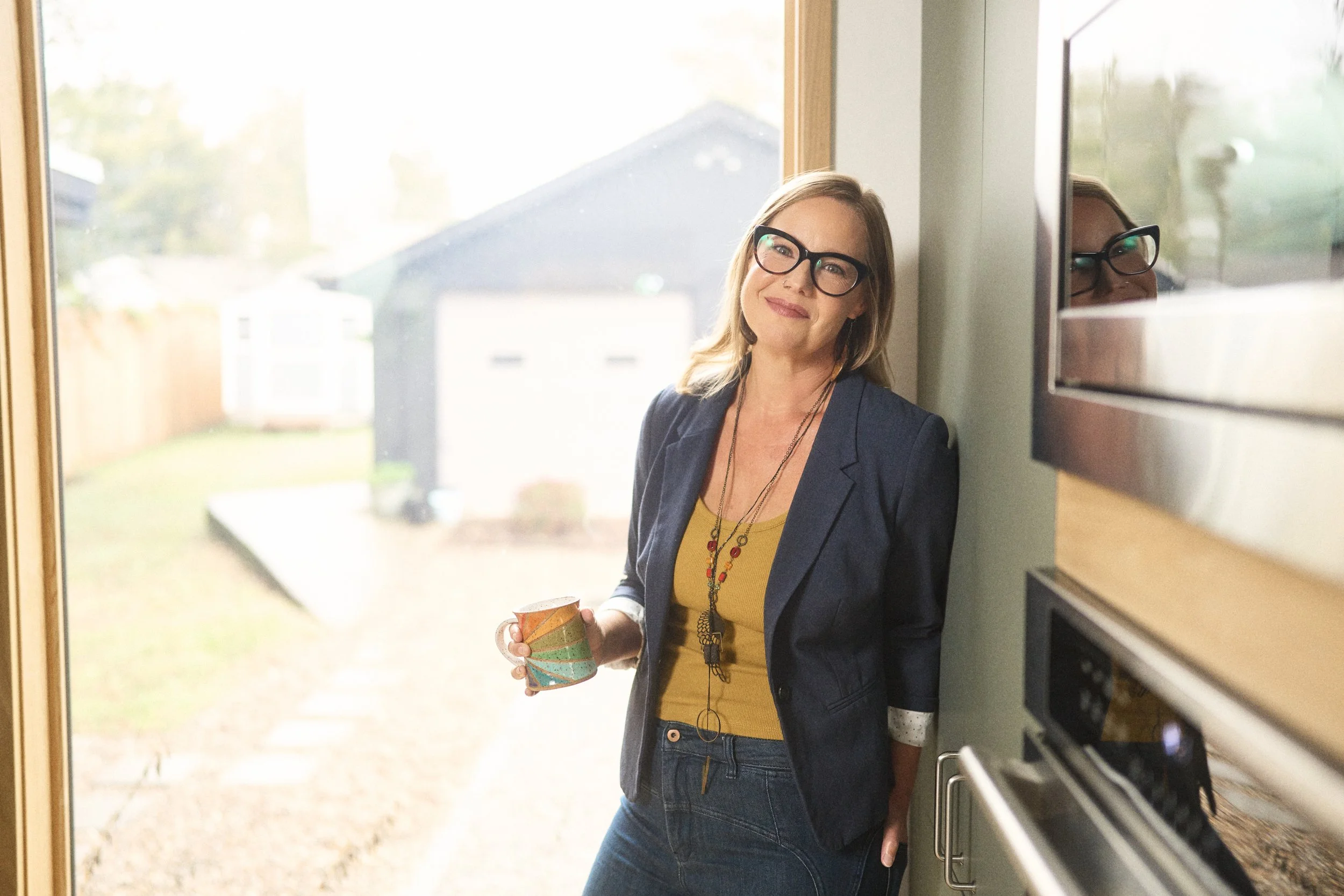 A woman with glasses standing in a doorway, holding a mug, smiling, and looking at the camera.