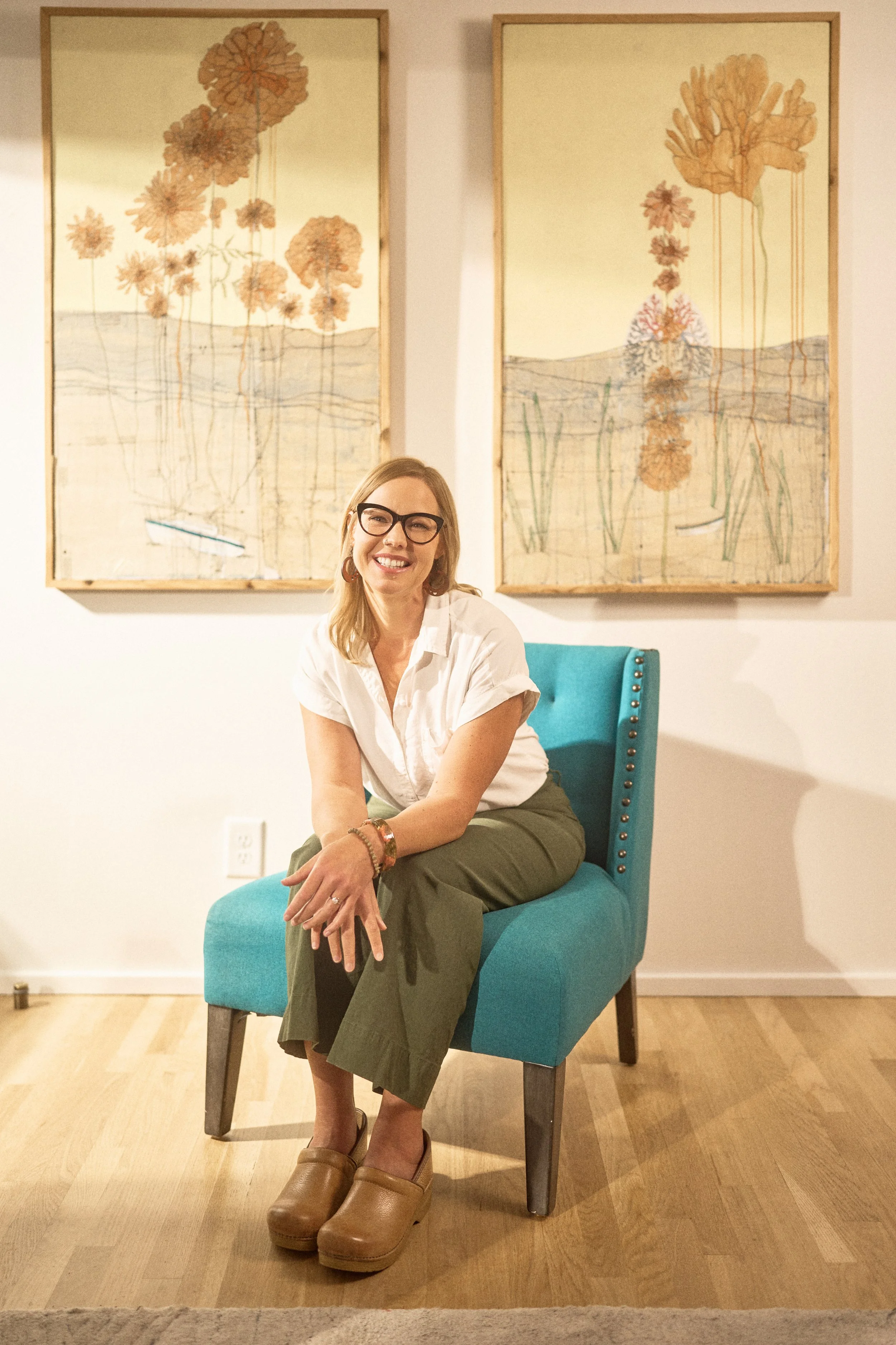 A woman with glasses, wearing a white shirt and green pants, sitting on a teal chair in front of two framed floral artwork pieces on a white wall.