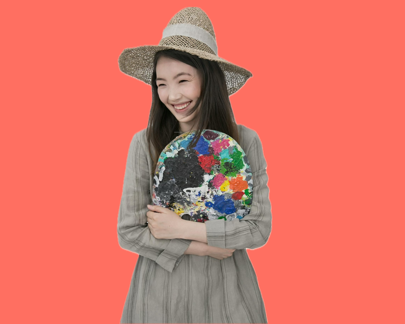 A smiling young woman wearing a large straw sun hat, holding a colorful painter's palette, standing against a coral background.