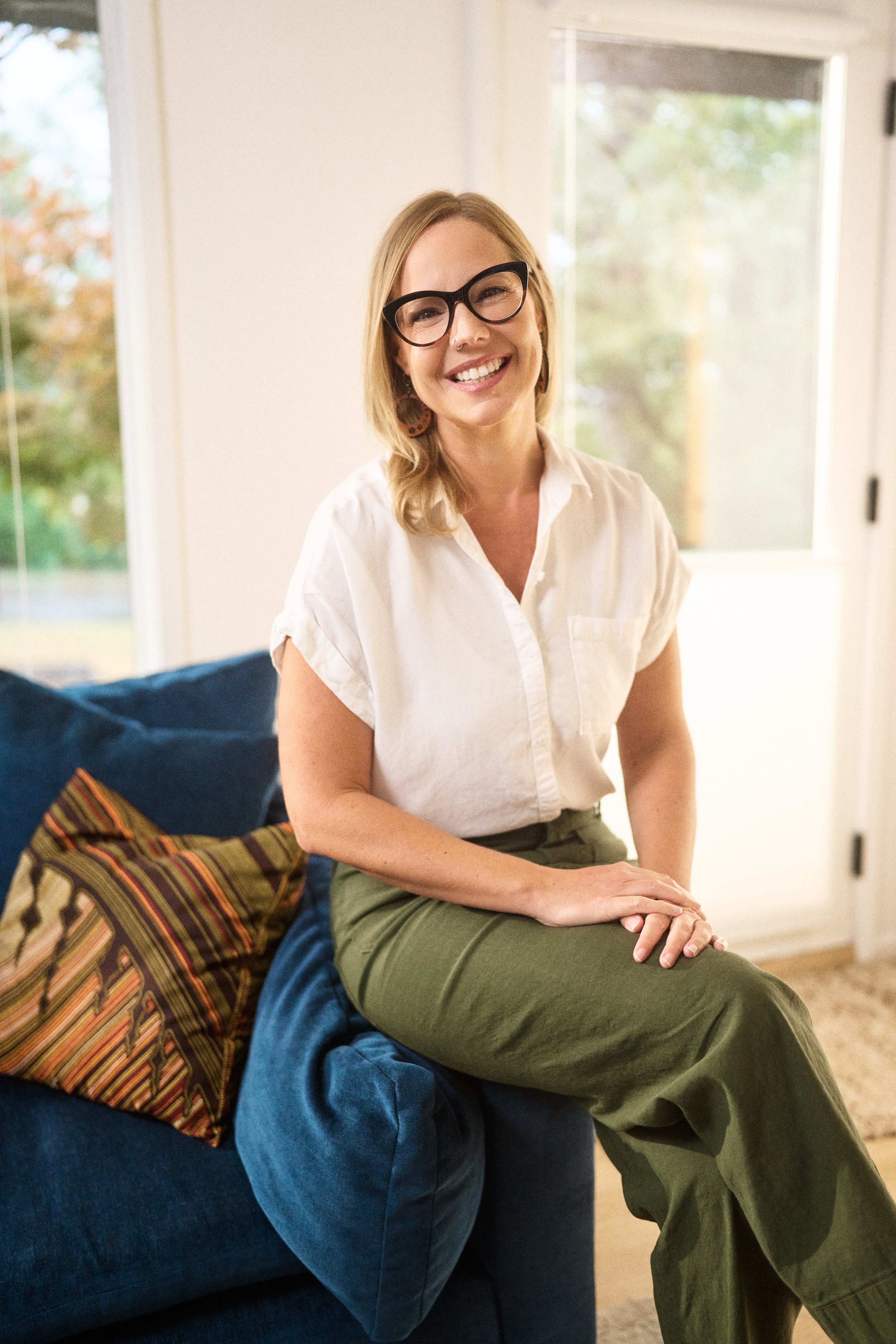 A woman with blonde hair, wearing glasses, a white shirt, and green pants, sitting on a blue couch indoors, smiling at the camera with a window showing trees outside in the background.