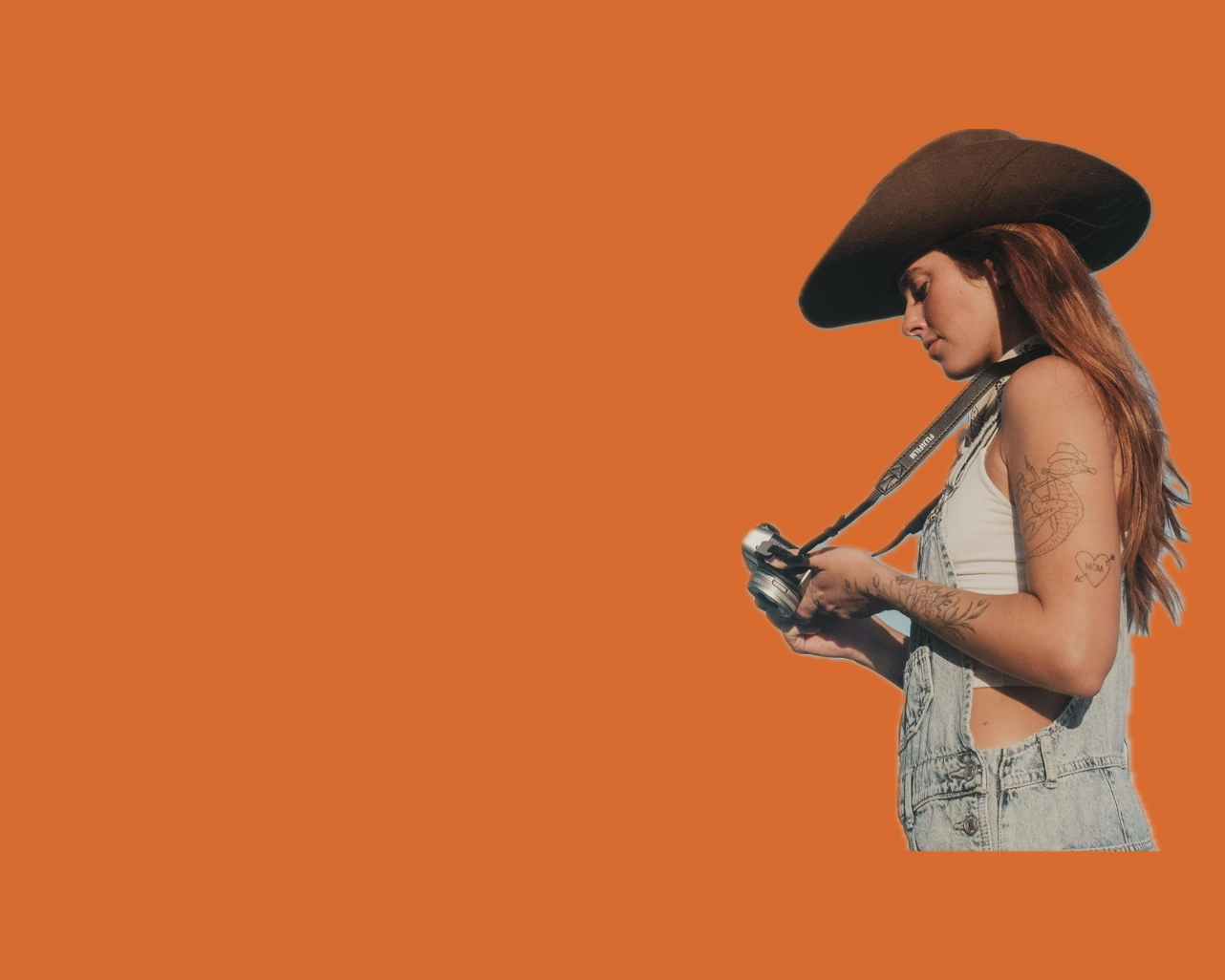 Woman with tattoos wearing a cowboy hat and denim vest, looking at camera, holding a camera with an orange background.