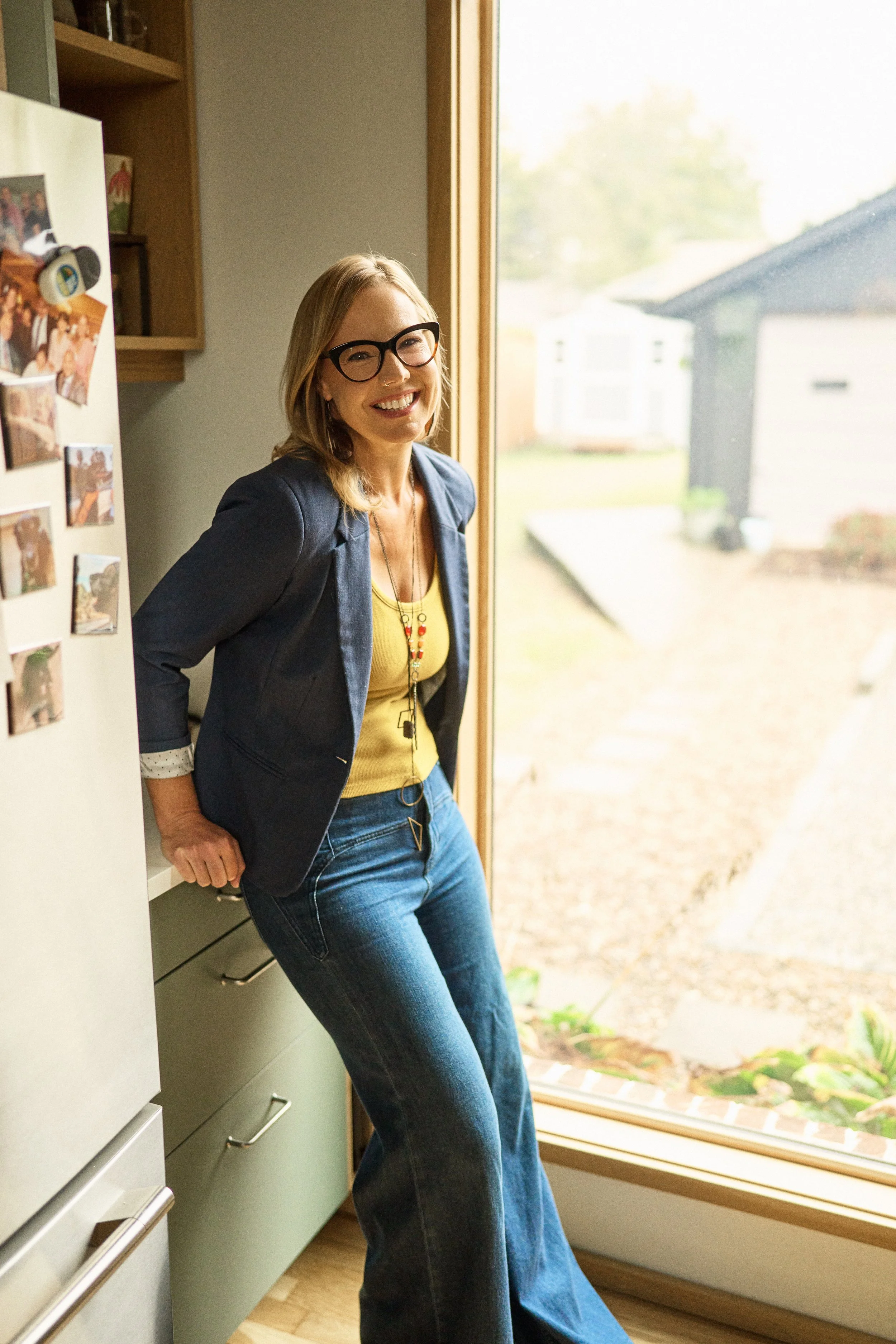 A woman with blonde hair, glasses, and a bright smile leaning next to a refrigerator in a kitchen, standing near a large window with a view of a backyard.
