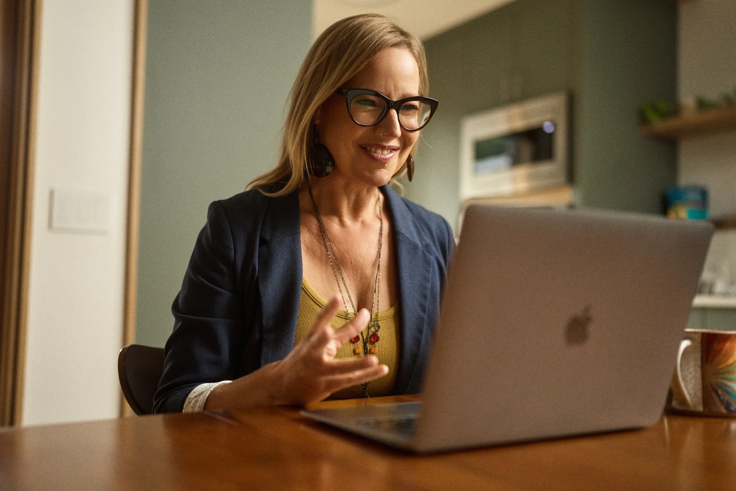Woman wearing glasses, a dark blazer, and a yellow top, sitting at a wooden table using a silver laptop, inside a kitchen or dining area.