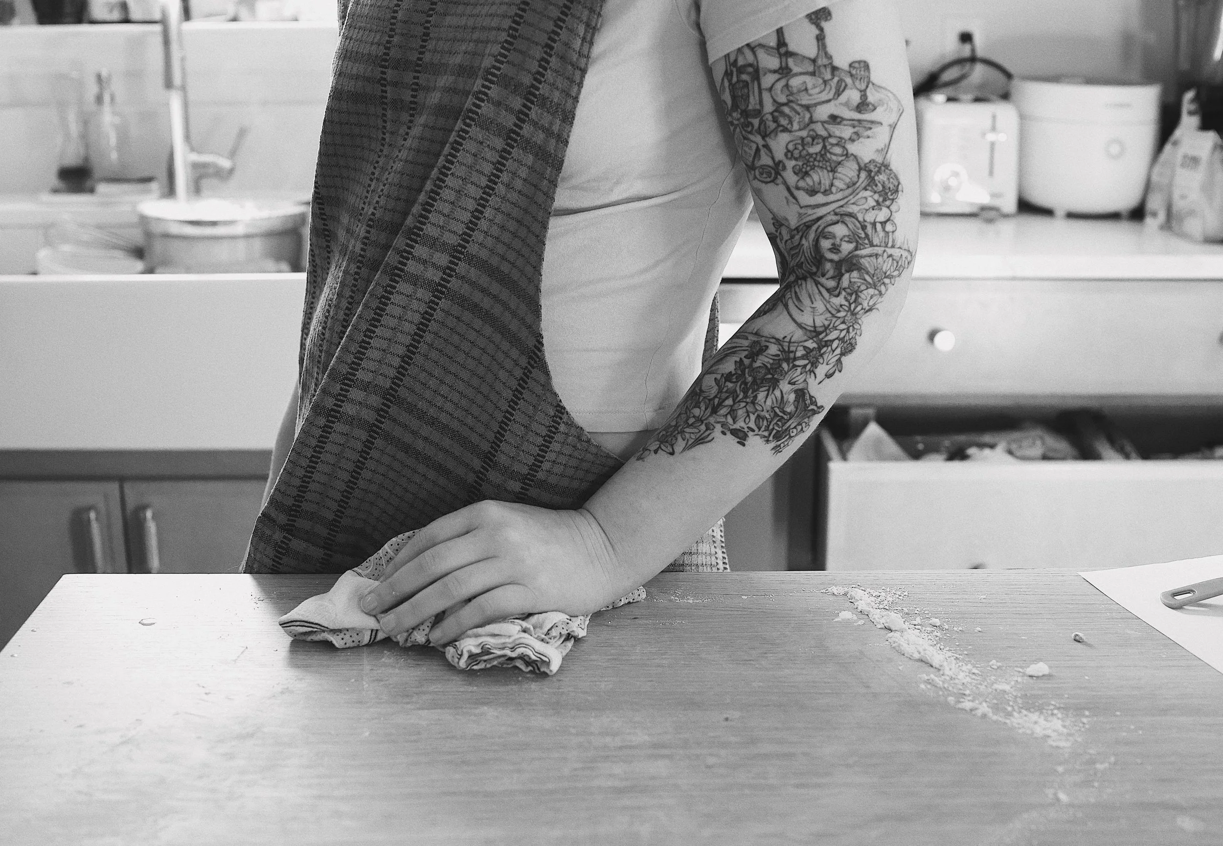Person with tattooed arm wiping a kitchen counter with a cloth, with flour and a rolling pin nearby.