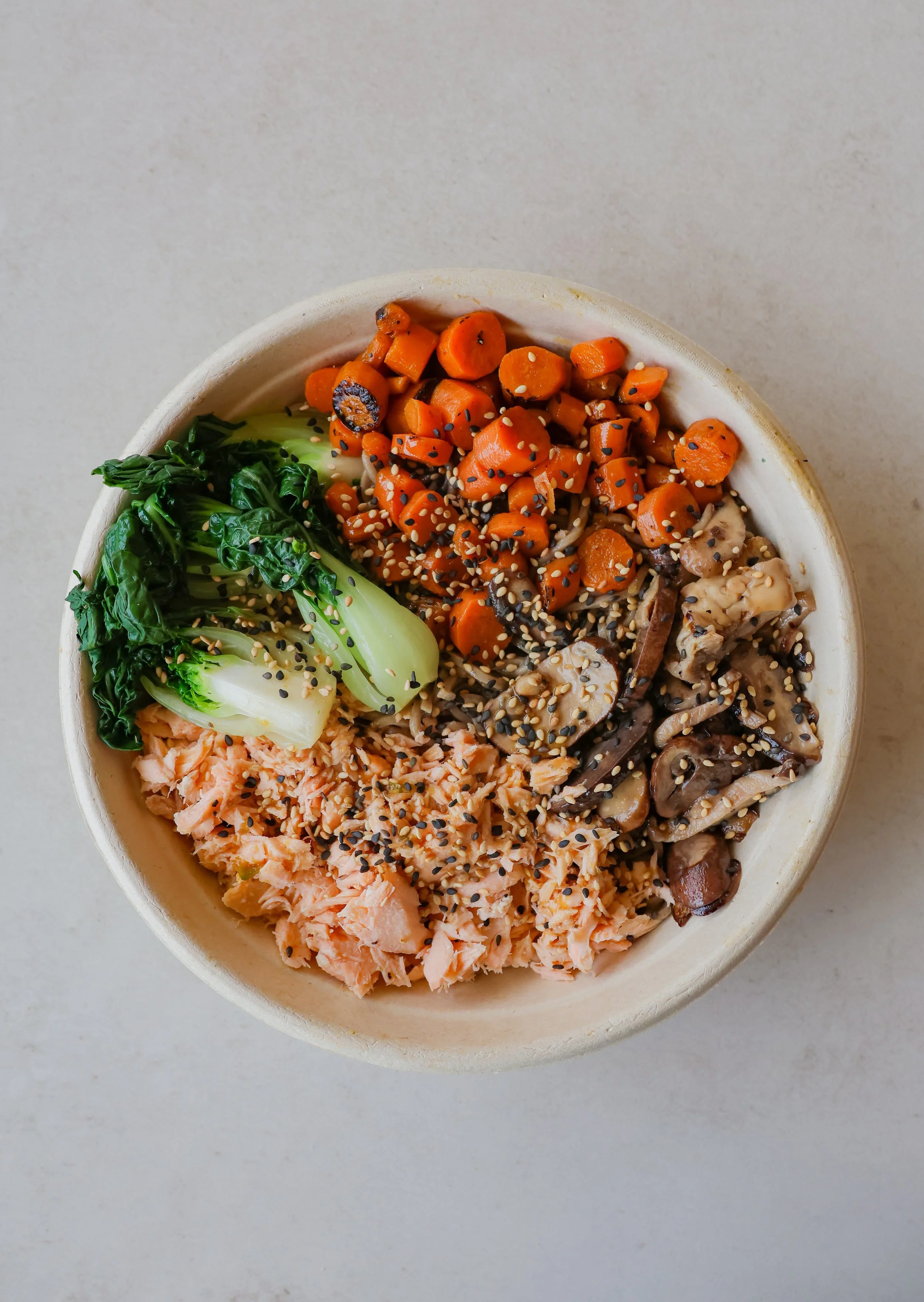 Bowl with assorted food ingredients including cooked carrots sprinkled with sesame seeds, greens, shredded salmon, and mushrooms, on a light surface.