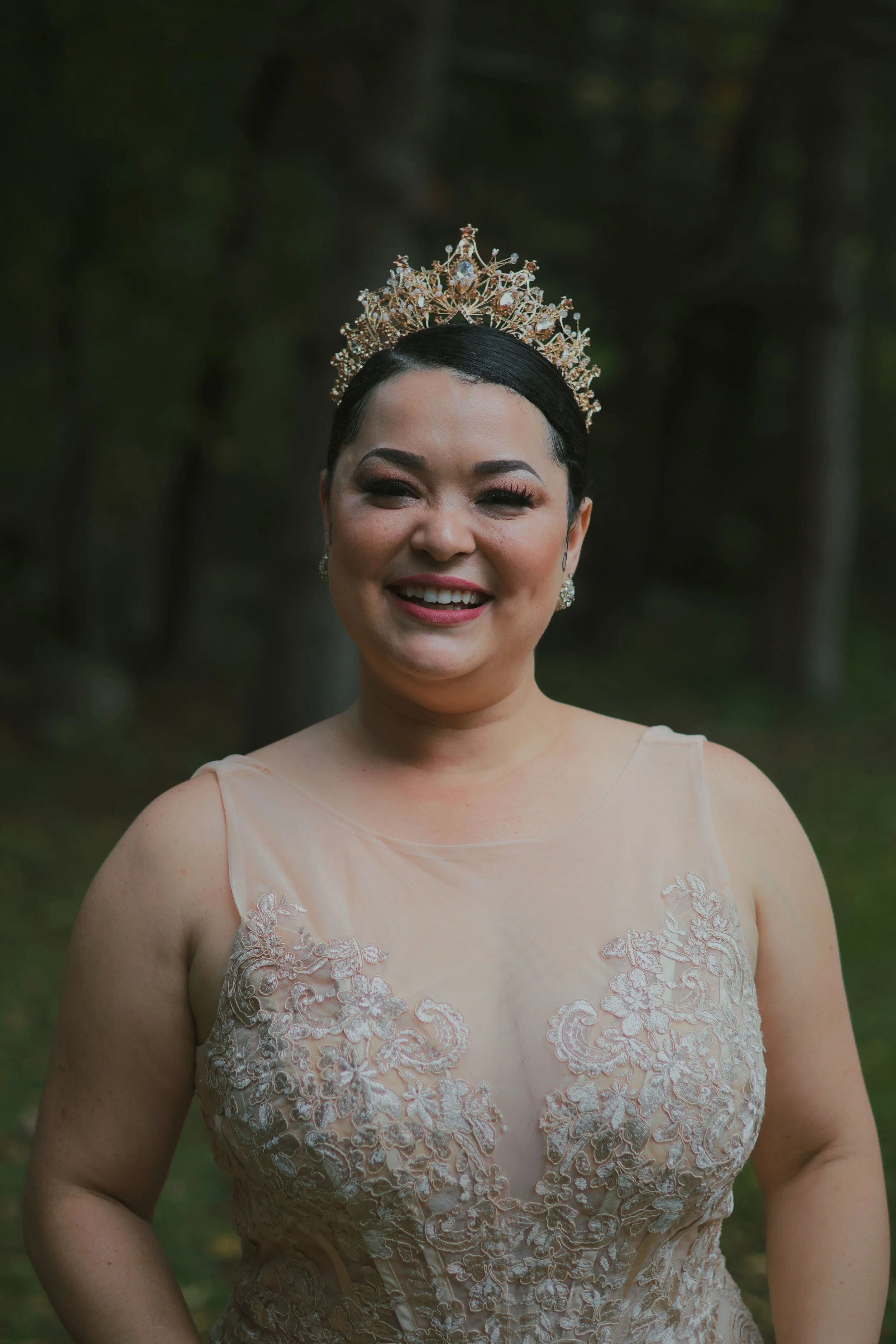 A woman with dark hair in an updo, wearing a tiara, earrings, and a sheer, embroidered dress, smiling outdoors in a forest setting.