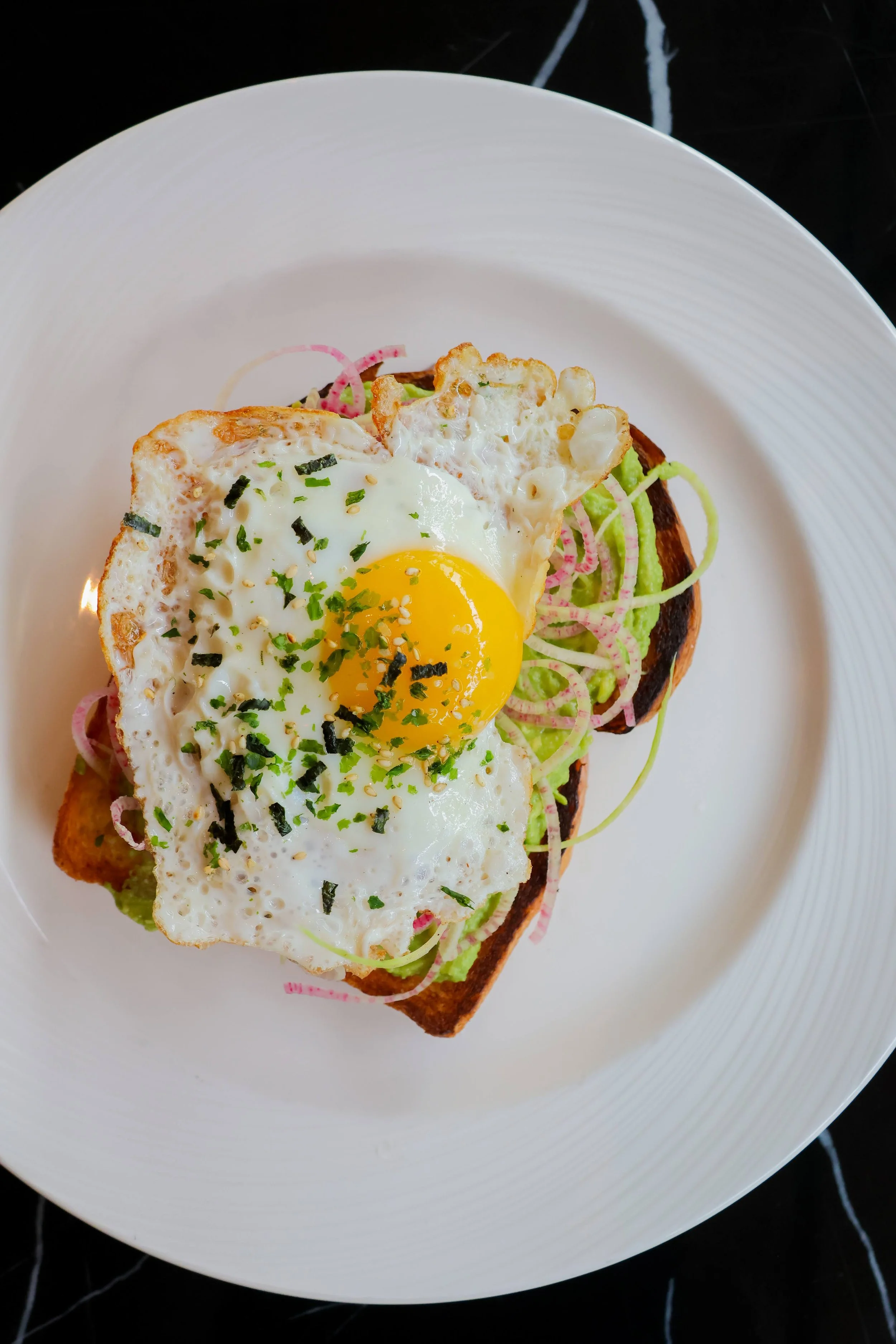 A plate with a piece of toast topped with a fried egg, chopped herbs, and black sesame seeds, served over a bed of sliced vegetables.