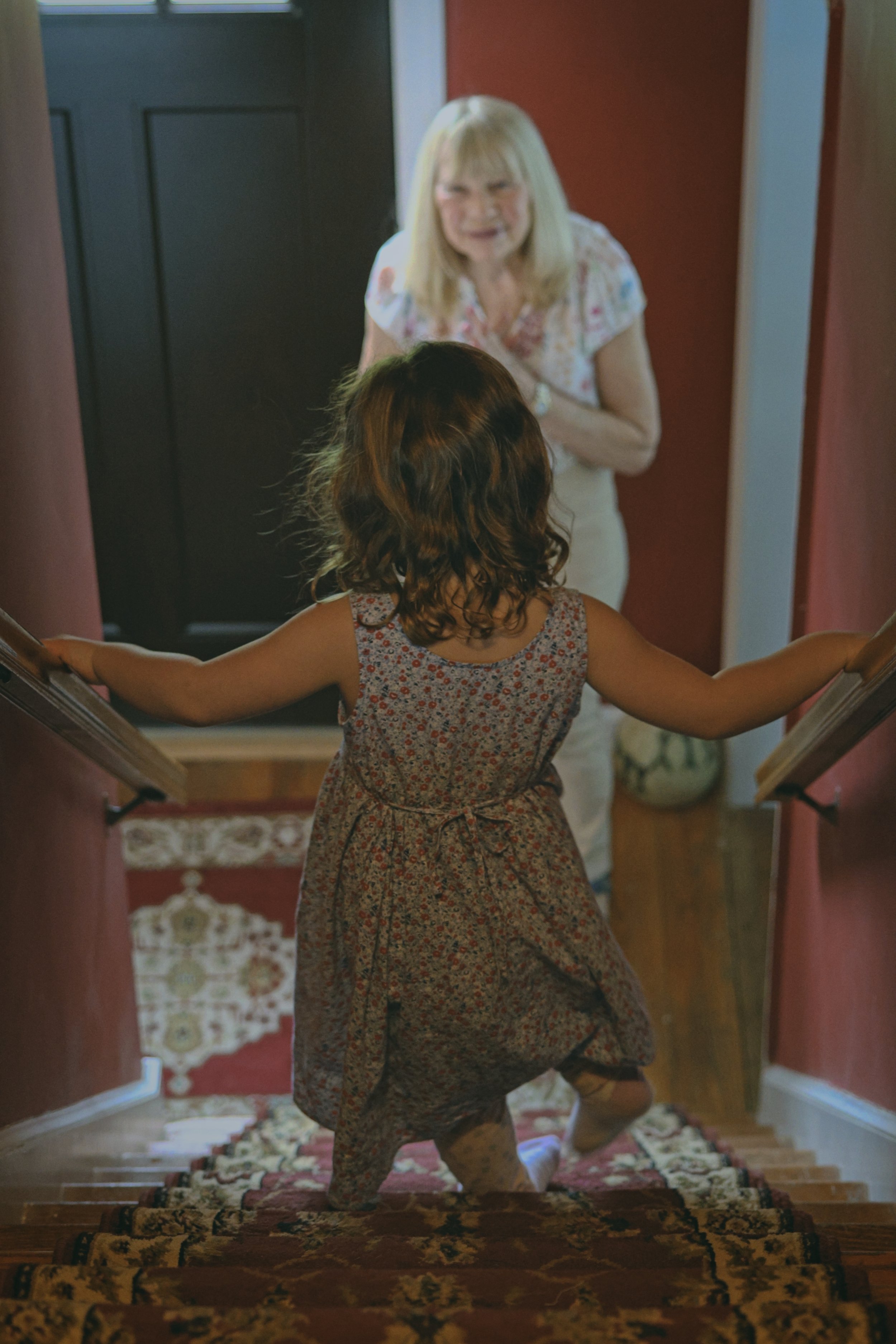 A young girl in a floral dress is running up a staircase with a floral carpet, while an older woman with blonde hair, wearing a light-colored dress, stands at the top of the staircase looking at her in Berkeley Heights, NJ.