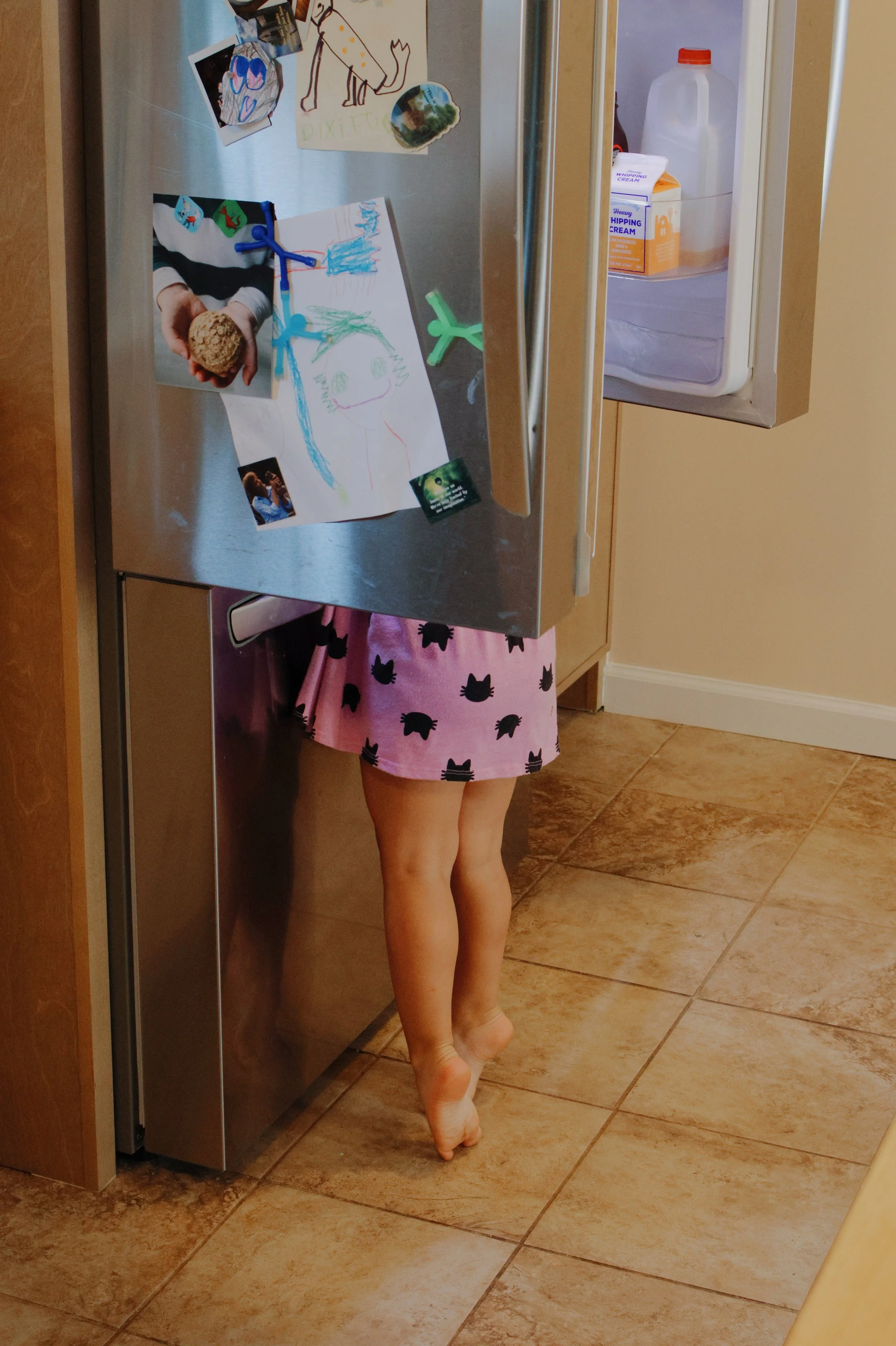 A child standing on the kitchen floor partially hidden behind a stainless steel refrigerator, with only their legs visible, wearing pink pajama shorts with black cat silhouettes.