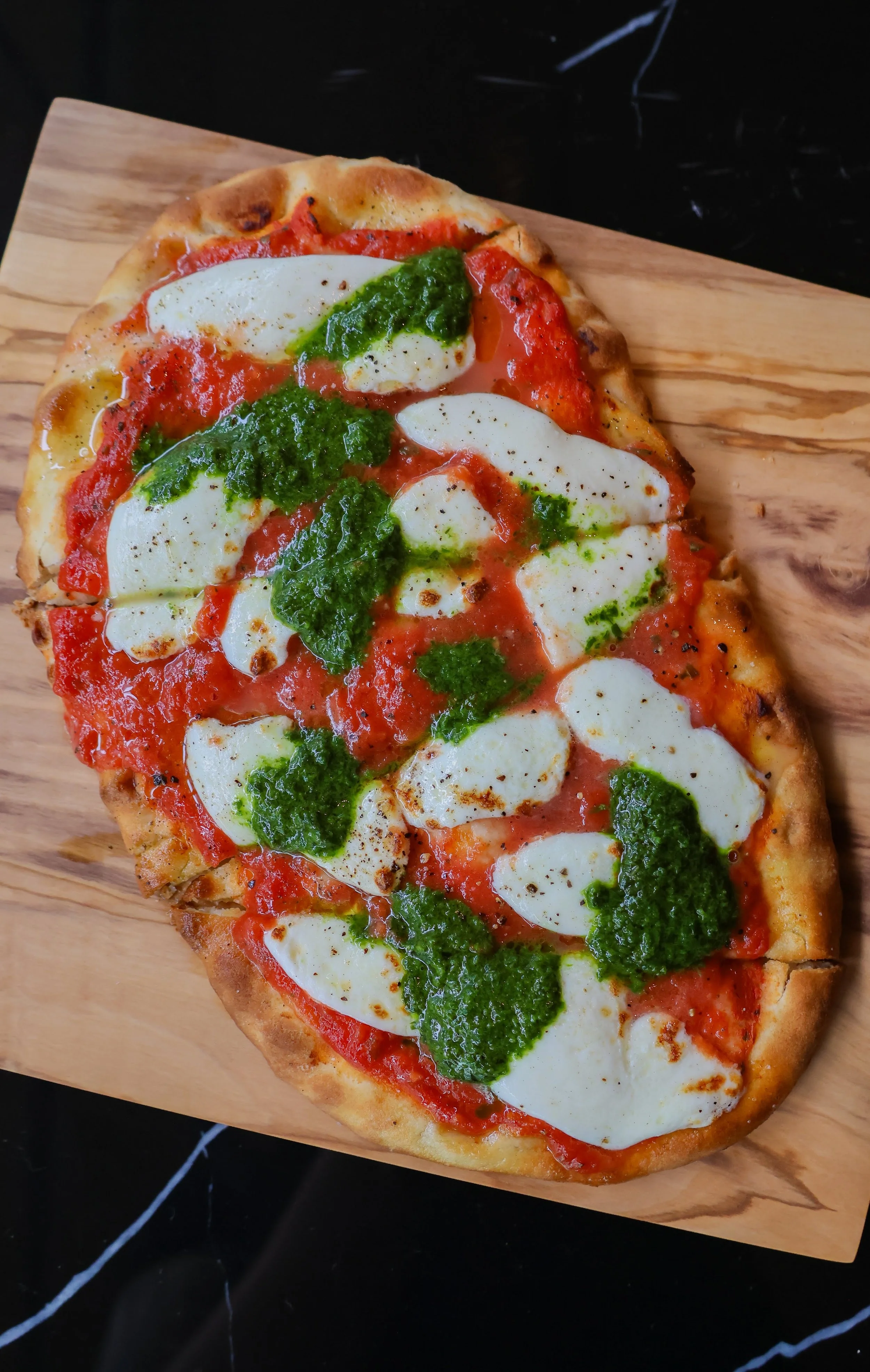 Pizza with cheese, tomato sauce, fresh mozzarella, basil pesto, and red bell peppers on a wooden serving board in Morristown, NJ. 