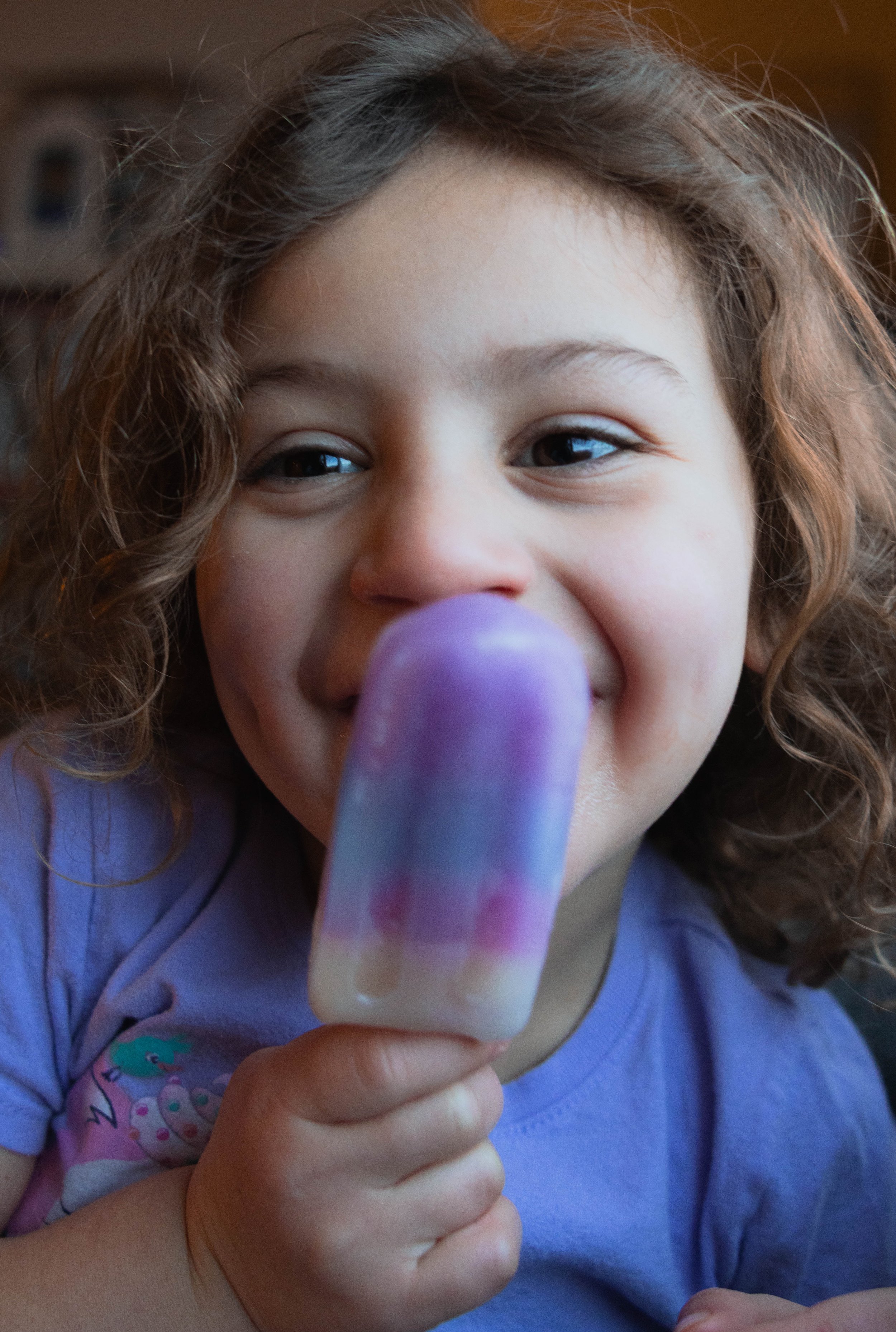 A young girl with curly hair is smiling and holding a colorful purple, pink, and cream popsicle close to her face.