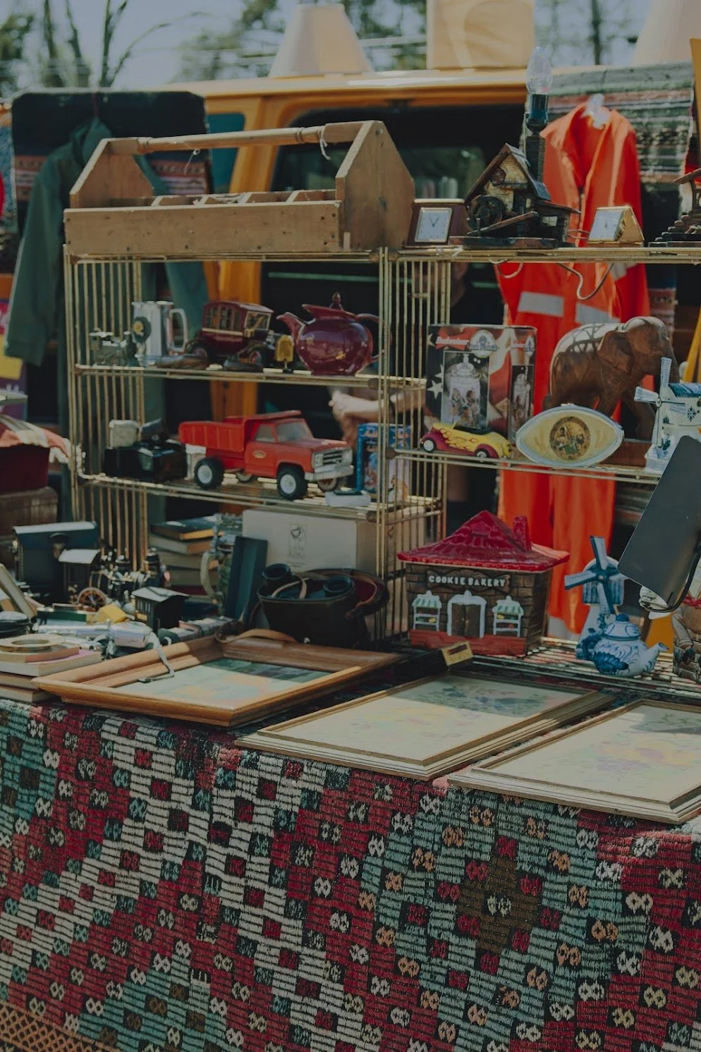 A flea market stall displaying various vintage toys, decorative items, and framed artwork on a table covered with a patterned cloth and a metal shelving unit, with a yellow vehicle in the background in Lambertville, NJ