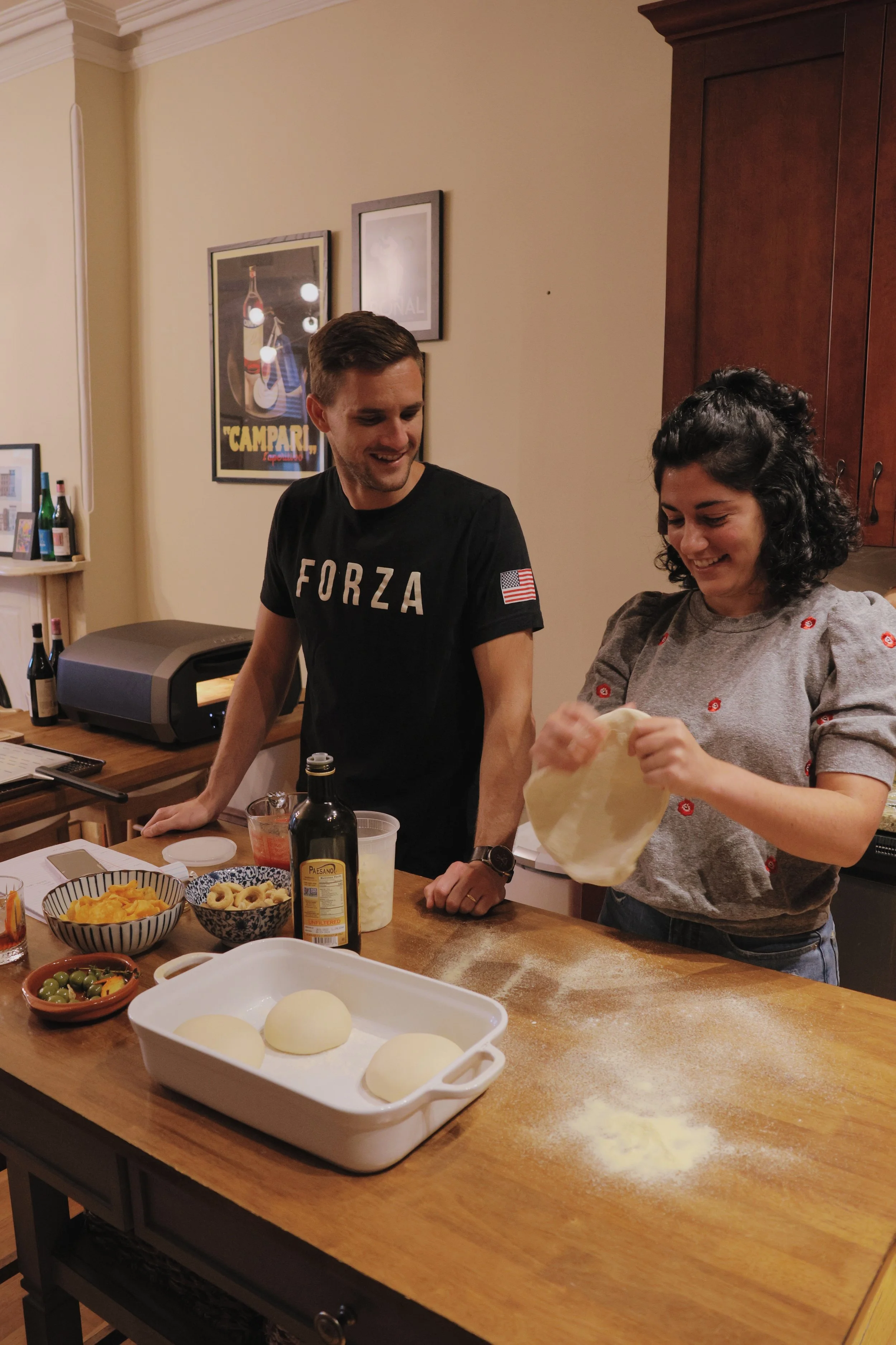 Two people are preparing pizza dough on a kitchen counter with various ingredients, including olives, cheese, and tomato sauce. One person is holding and working with the dough, smiling, while the other stands nearby also smiling. The kitchen has fra