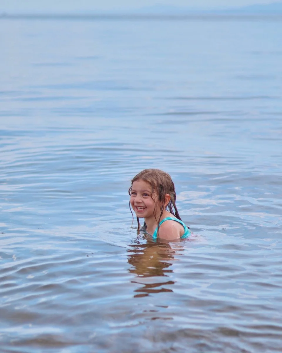 A young girl with wavy hair swimming in a body of water, smiling and looking to her left, with the water reaching her shoulders.