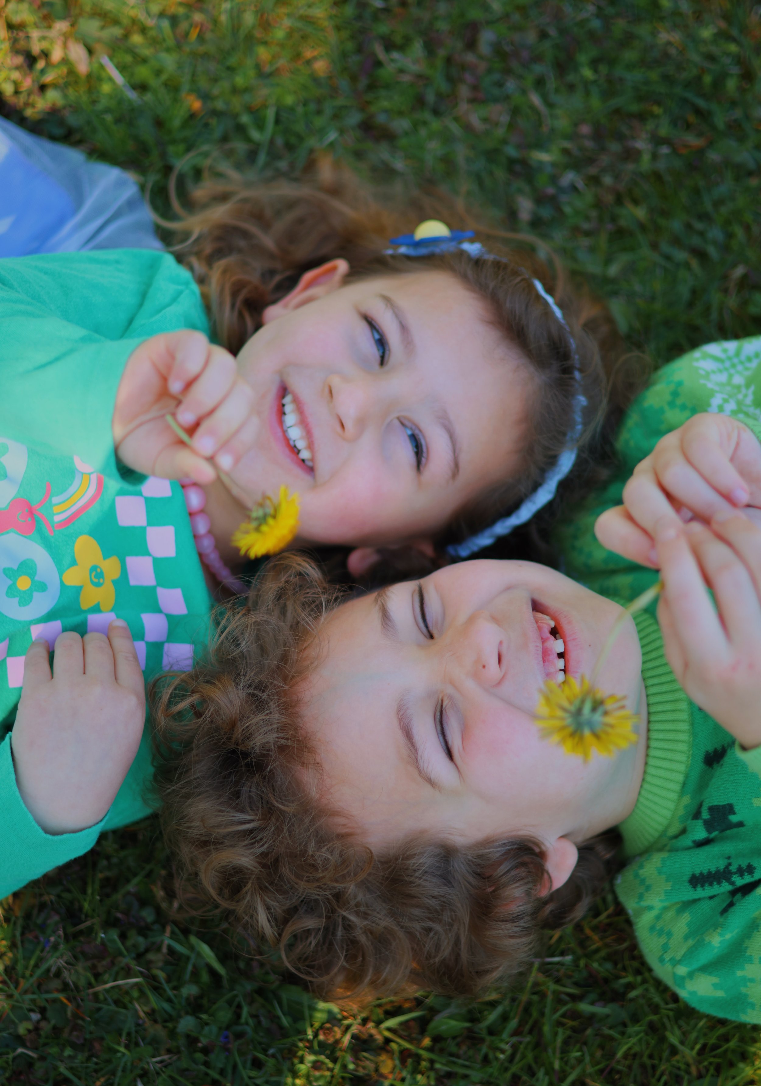 Two young girls lying on grass outdoors, smiling and holding yellow dandelions, enjoying a sunny day.