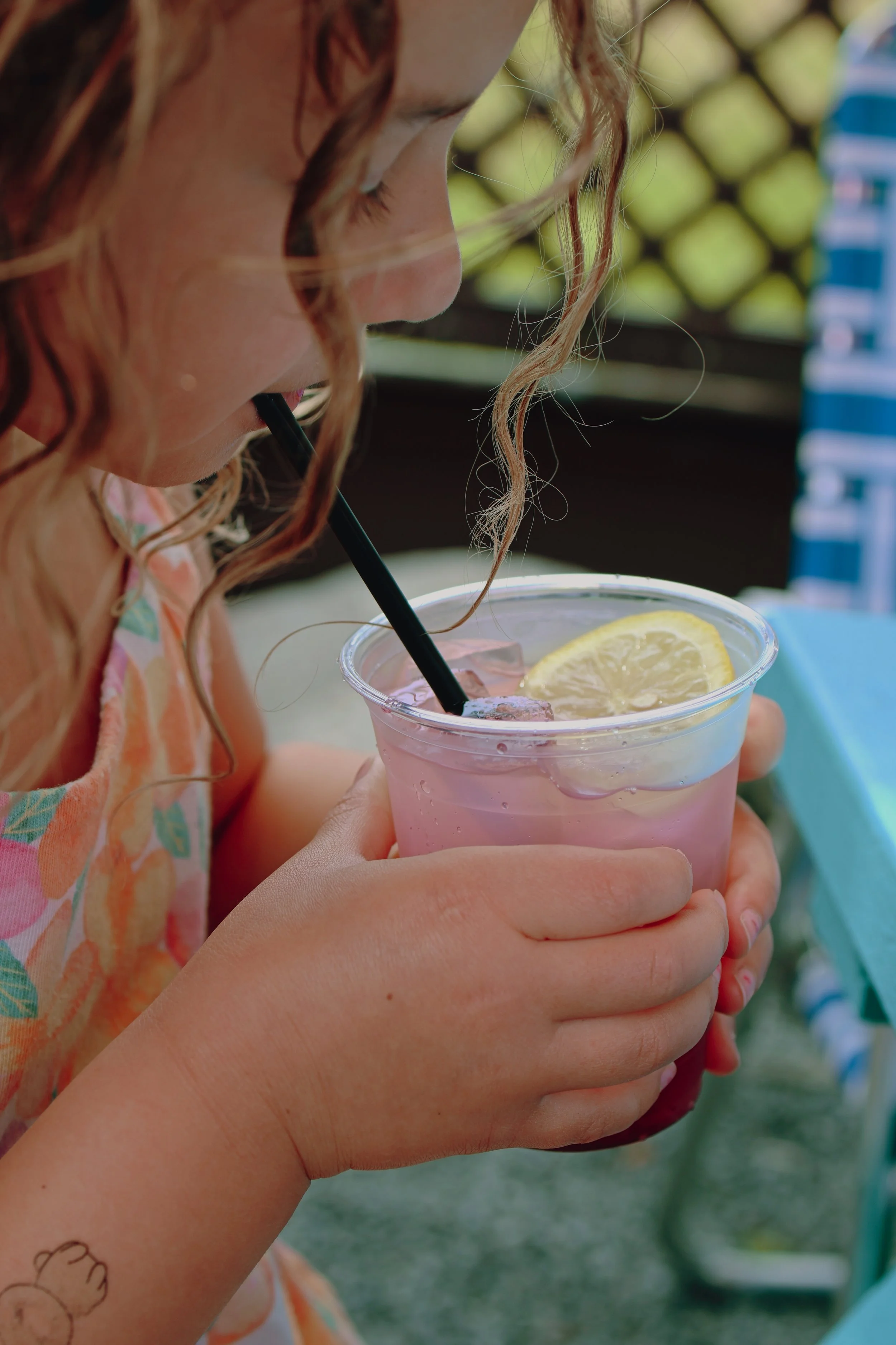 A young girl with curly hair drinking blueberry lemonade with lemon slice garnish through a black straw, holding the cup with both hands outdoors in Asbury Park, NJ at Porta.