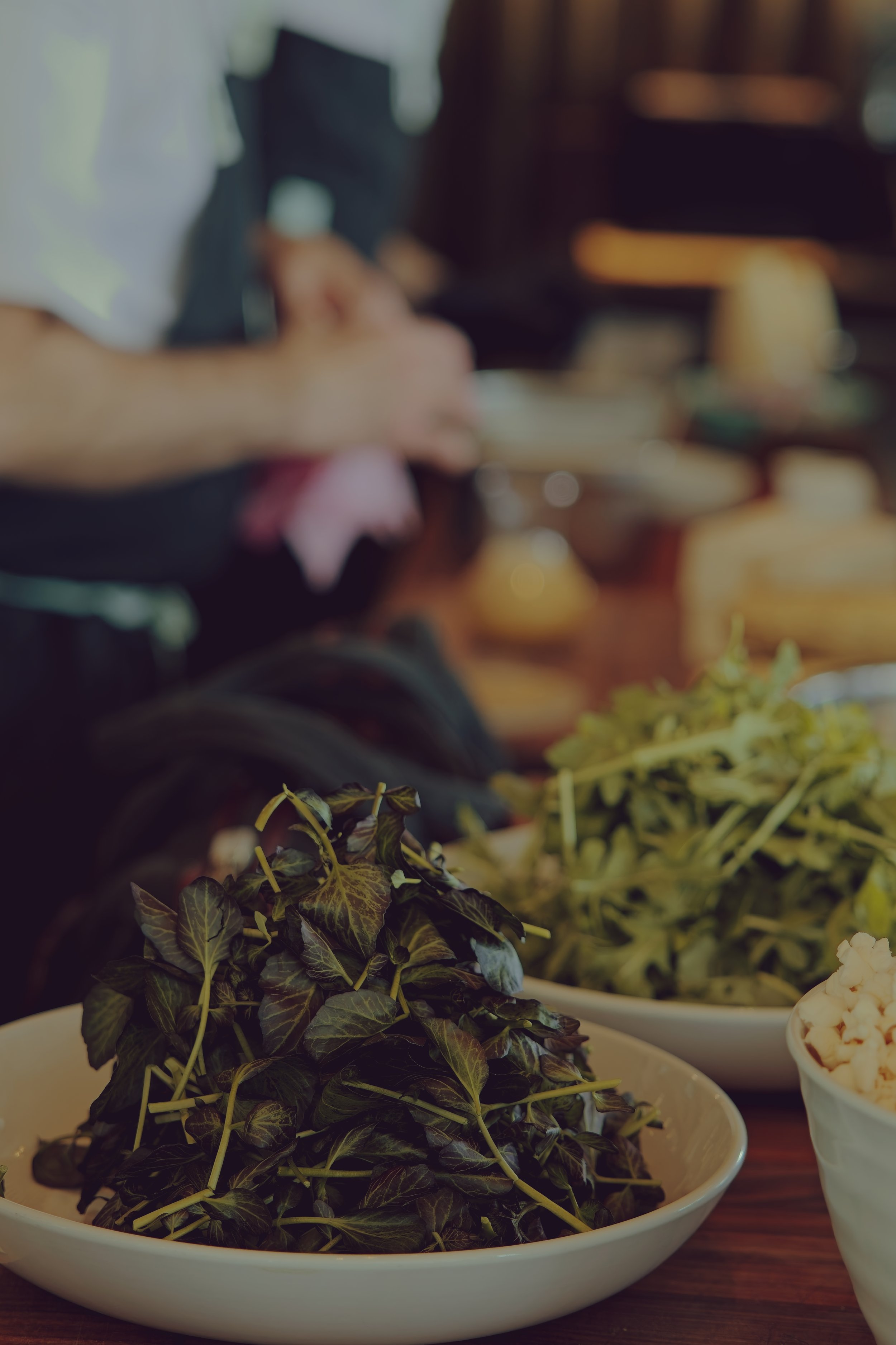 Close-up of a bowl filled with fresh green herbs, with other bowls of herbs and food items in the background on a dining table.