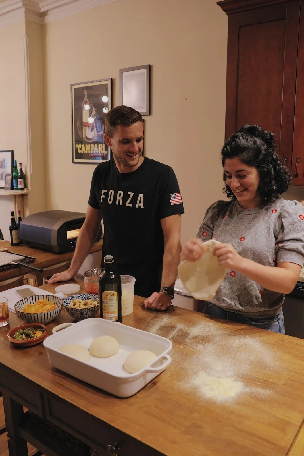 A man and woman happily preparing pizza dough in a kitchen, with various ingredients and a baking tray on the table.