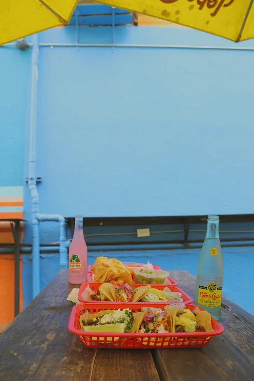 Outdoor table with three red plastic baskets filled with tacos, two glass bottles of flavored water, and a pink flavored soda bottle, with a yellow umbrella overhead and a light blue wall in the background.