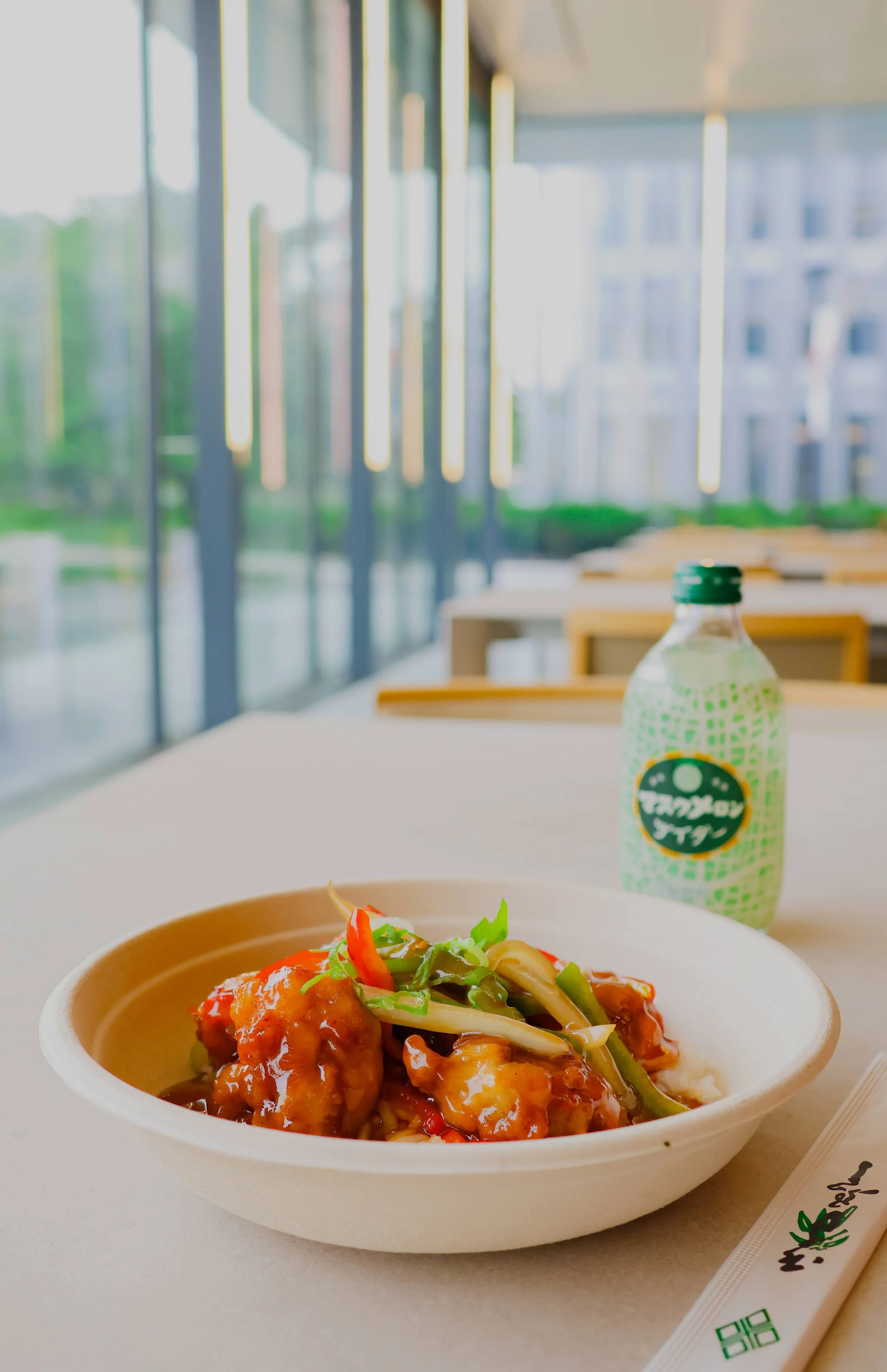 A bowl of Asian-style food with fried items, vegetables, and sauce, on a white table inside a modern, windowed restaurant with a blurred cityscape view outside. A bottled beverage and chopsticks are also on the table.