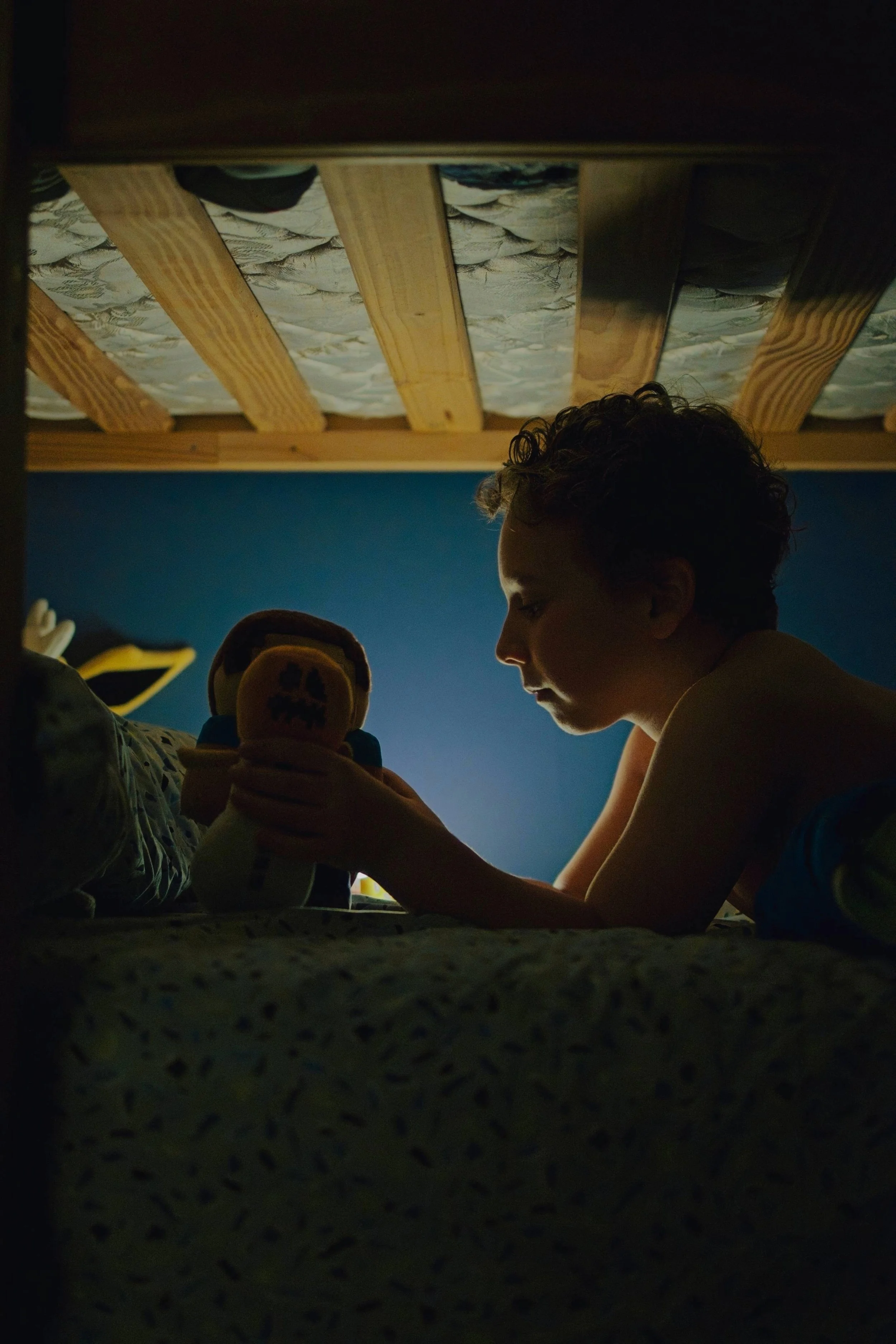 A young boy with curly hair sitting on a bed, illuminated by the glow of a device or toy he's holding, with a dark blue wall behind him and a bed with a wooden frame and patterned mattress.