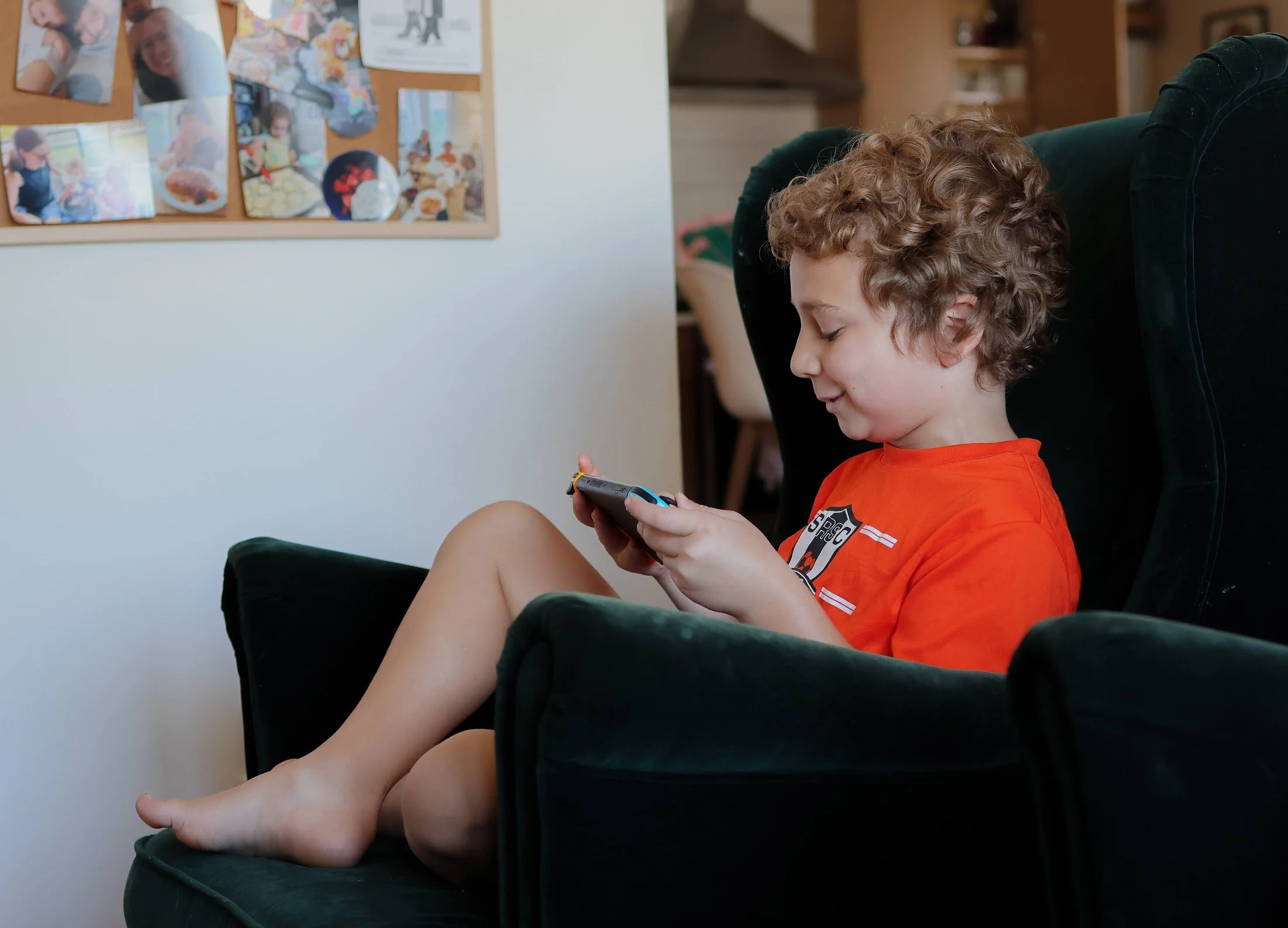 A young boy with curly hair in an orange shirt sitting in a green armchair, looking at a handheld gaming device.