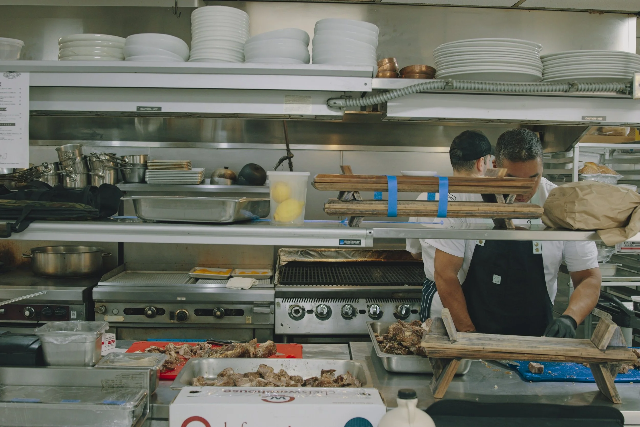 Documentary photo of a commercial kitchen with two chefs preparing foodm surrounded by plates, pots, and kitchen utensils.