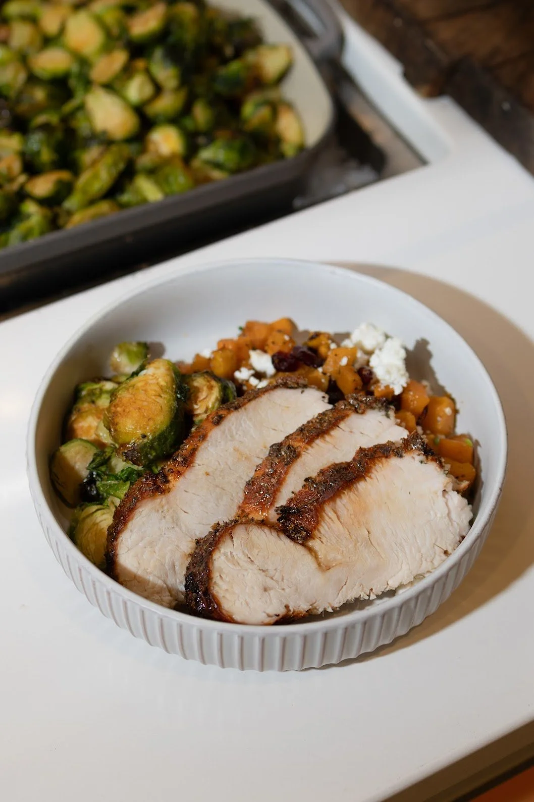 A white bowl filled with sliced roasted chicken breast, roasted Brussels sprouts, and a mixture of sweet potatoes, cranberries, and crumbled blue cheese.