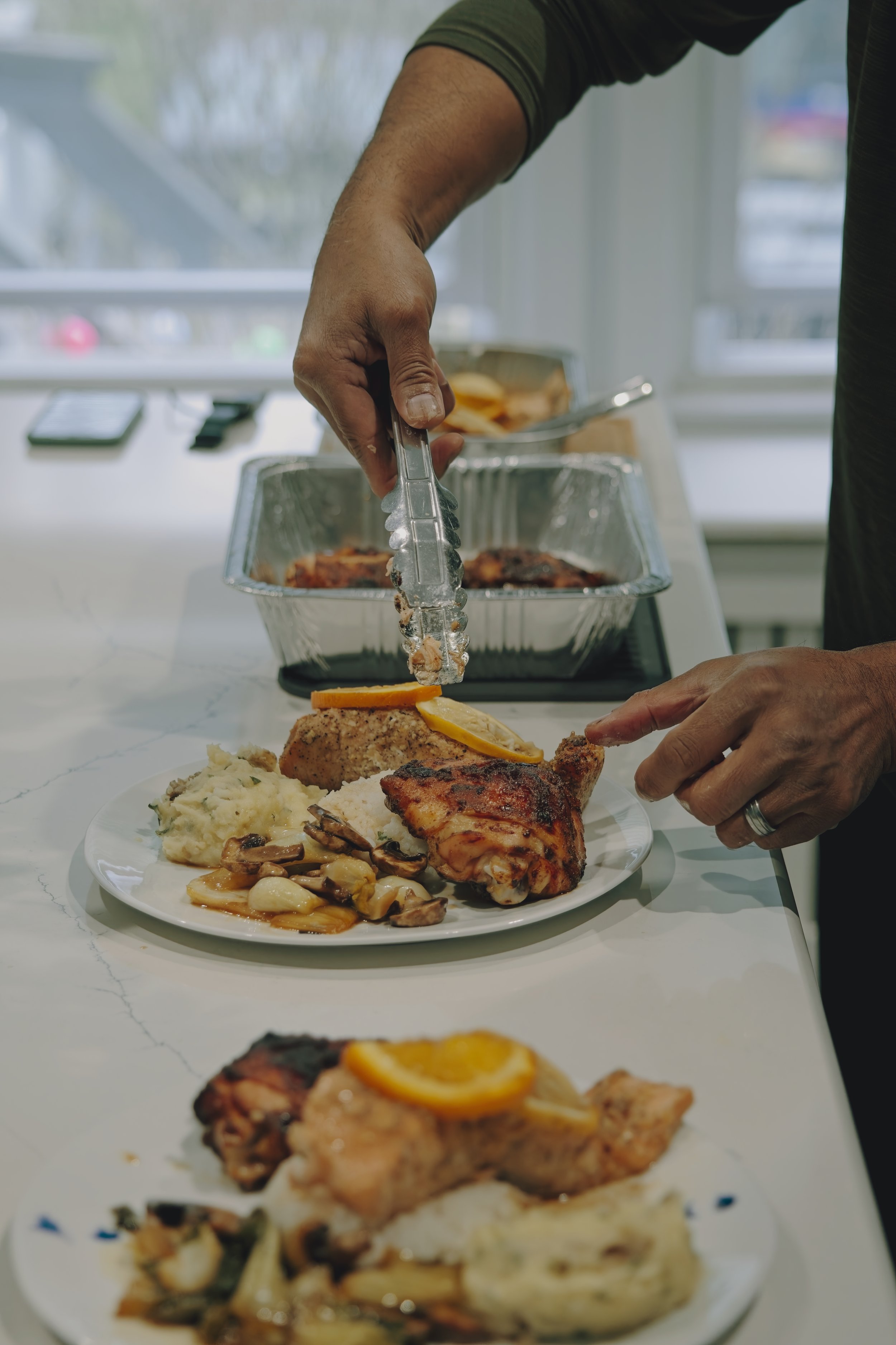 A person is serving food onto a plate that has mashed potatoes, chicken, and vegetables. There are aluminum containers with more food in the background, and the person is using tongs to place more food on the plate. New Providence, NJ. 