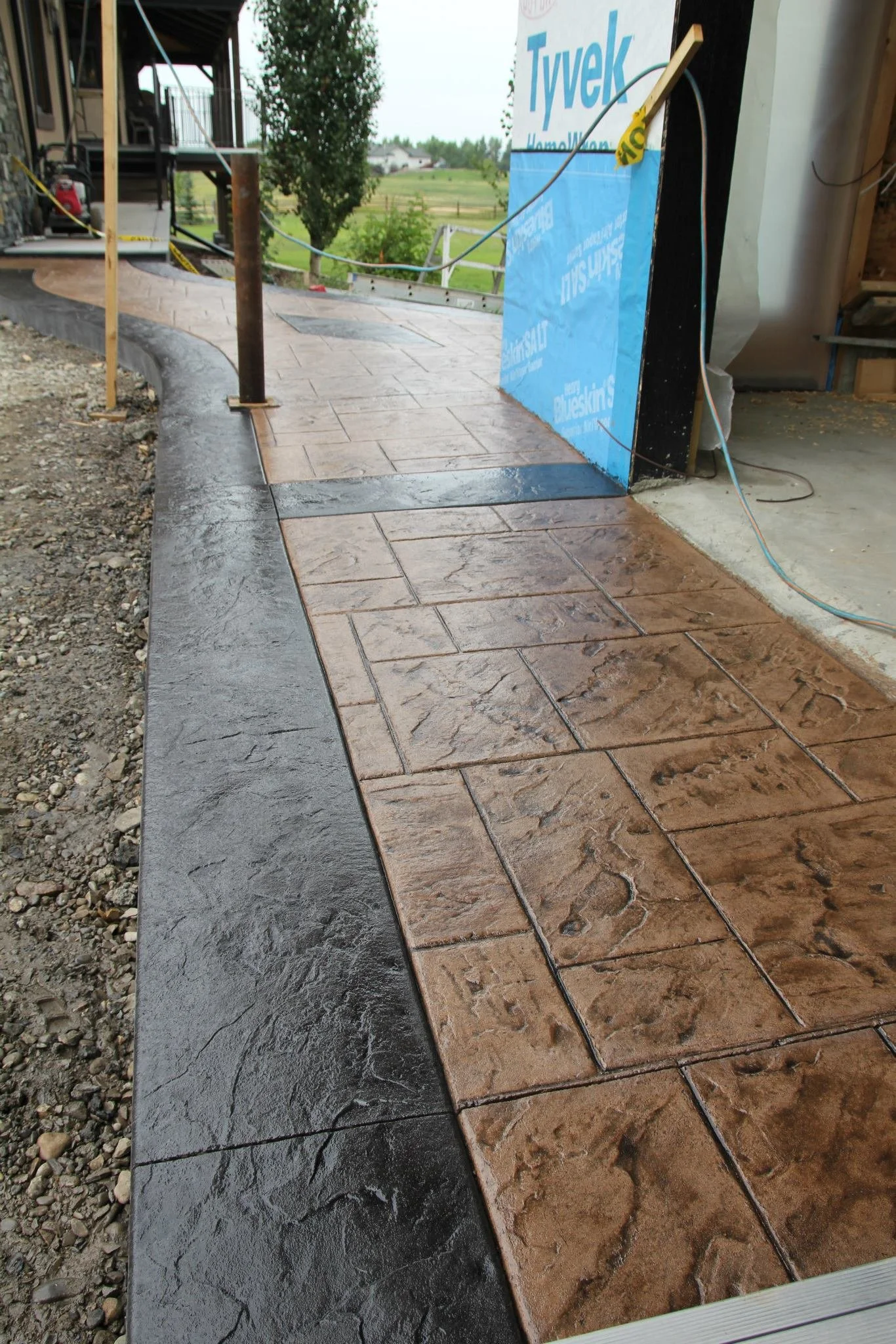 Freshly installed stamped concrete walkway with a black border adjacent to a house under construction, featuring a Tyvek house wrap on the wall.