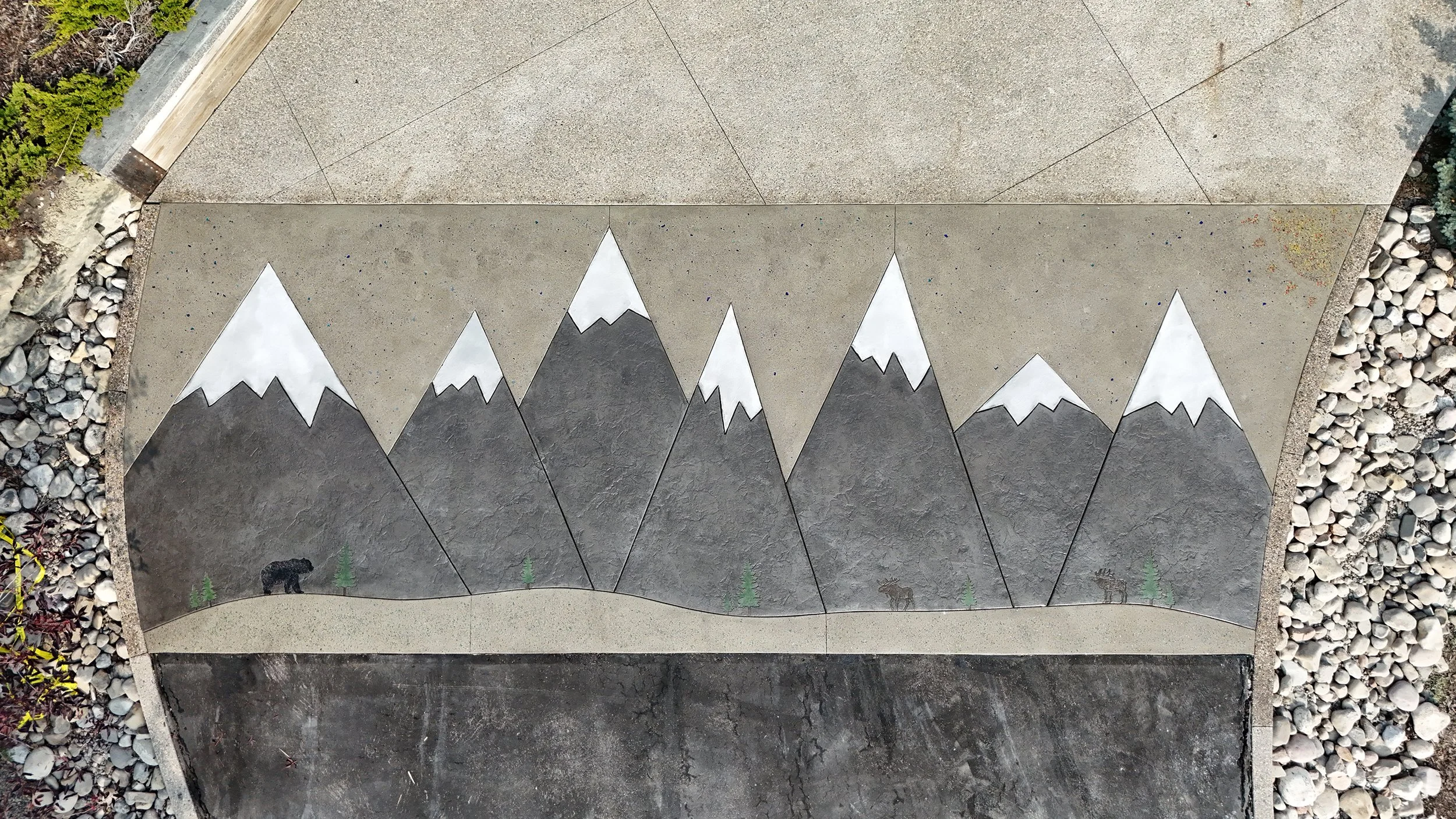 Outdoor mural of a mountain range with white-capped peaks, green pine trees, a black bear, a moose, and a landscape background, painted on concrete paving stones.