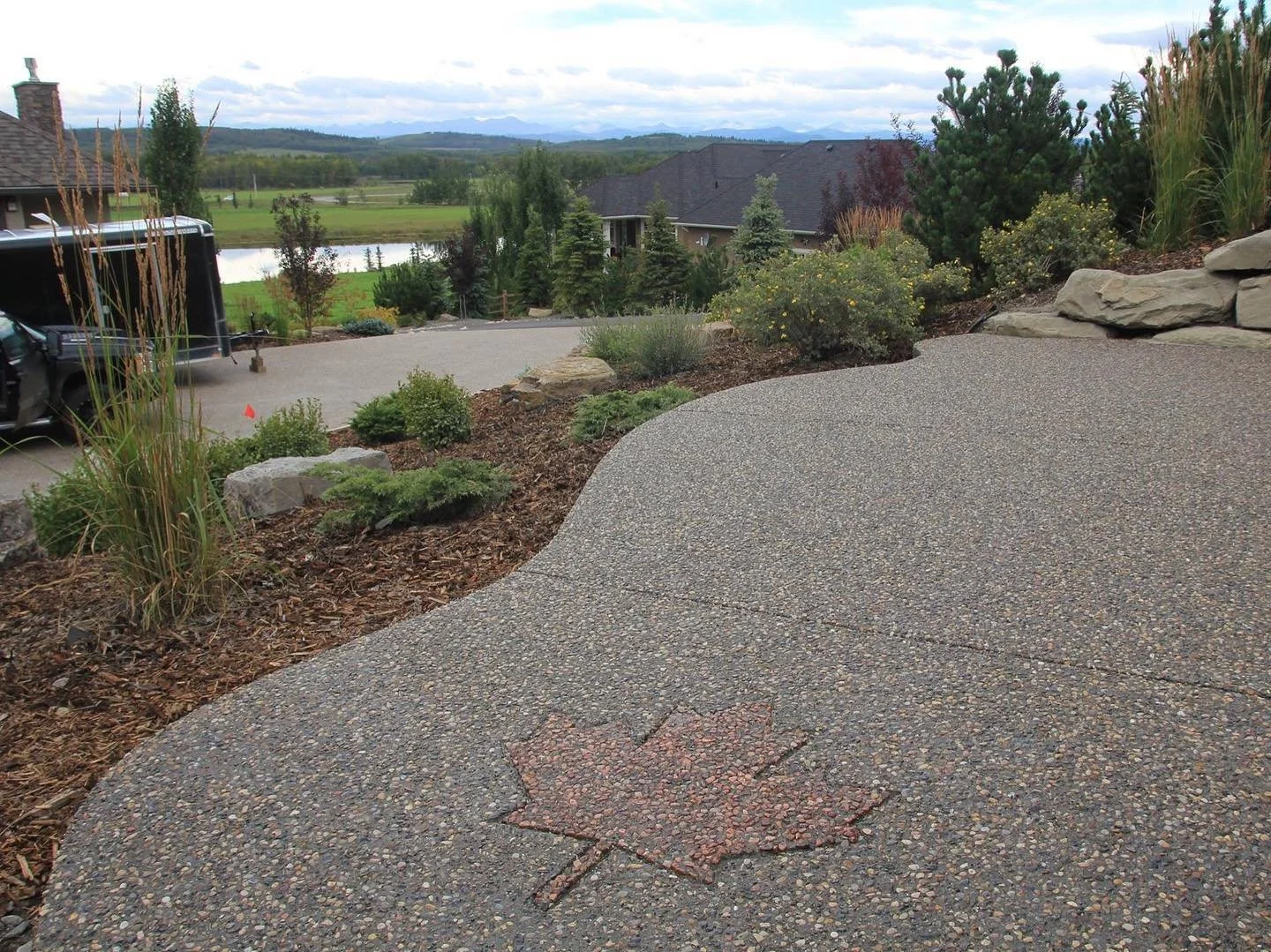 A paved outdoor area with a decorative maple leaf design embedded in the concrete, surrounded by landscaped garden beds and trees, overlooking a body of water and distant mountains.