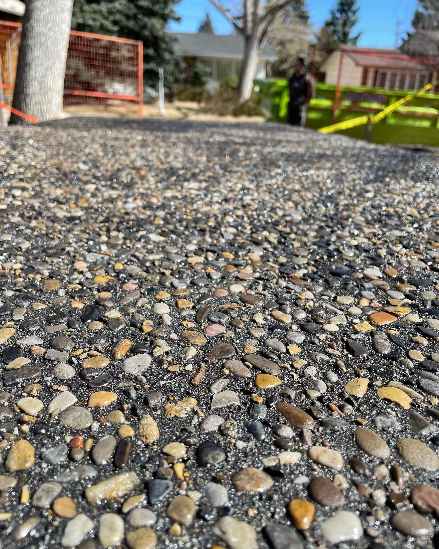 Close-up of a rough asphalt sidewalk with small multicolored stones, in a residential neighborhood with trees, houses, and a person in the background.