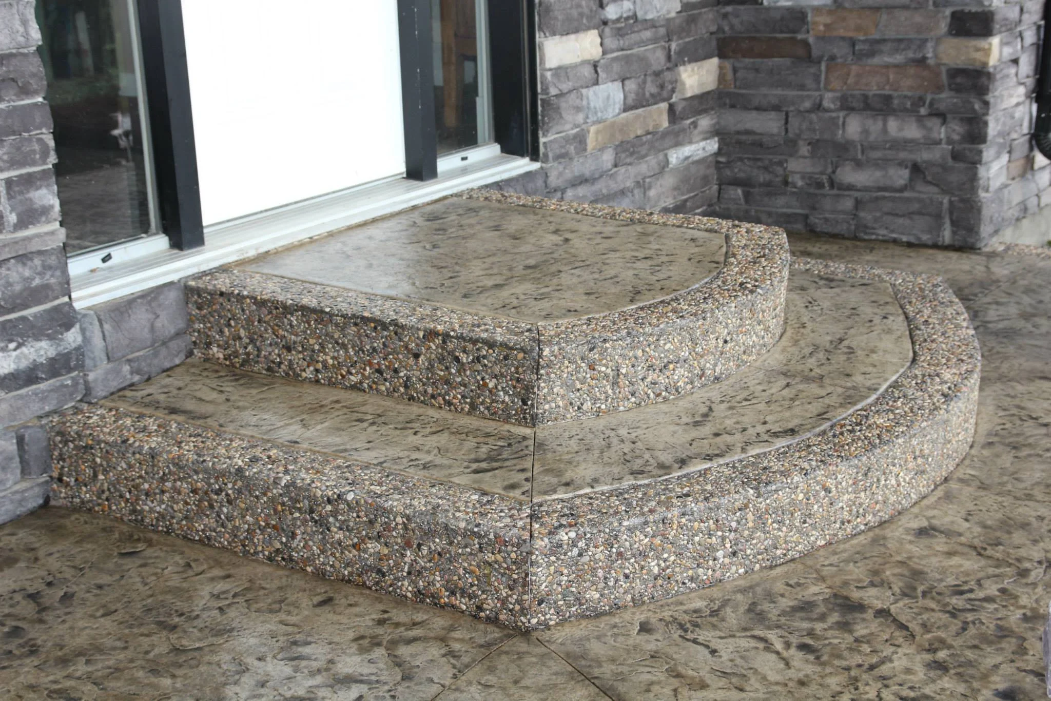 Two concrete steps with rounded edges leading to a glass door, surrounded by a brick and stone wall.