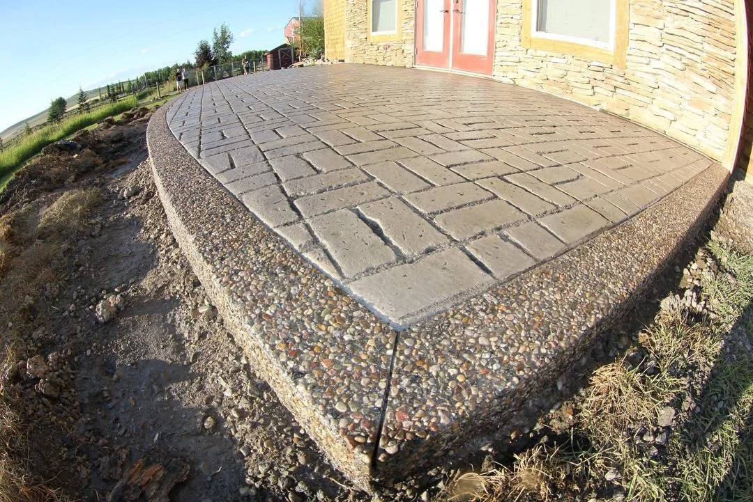 A newly constructed front porch with a brick pattern and a gravel border, attached to a brick house with a red door and two windows, in a rural outdoor setting.